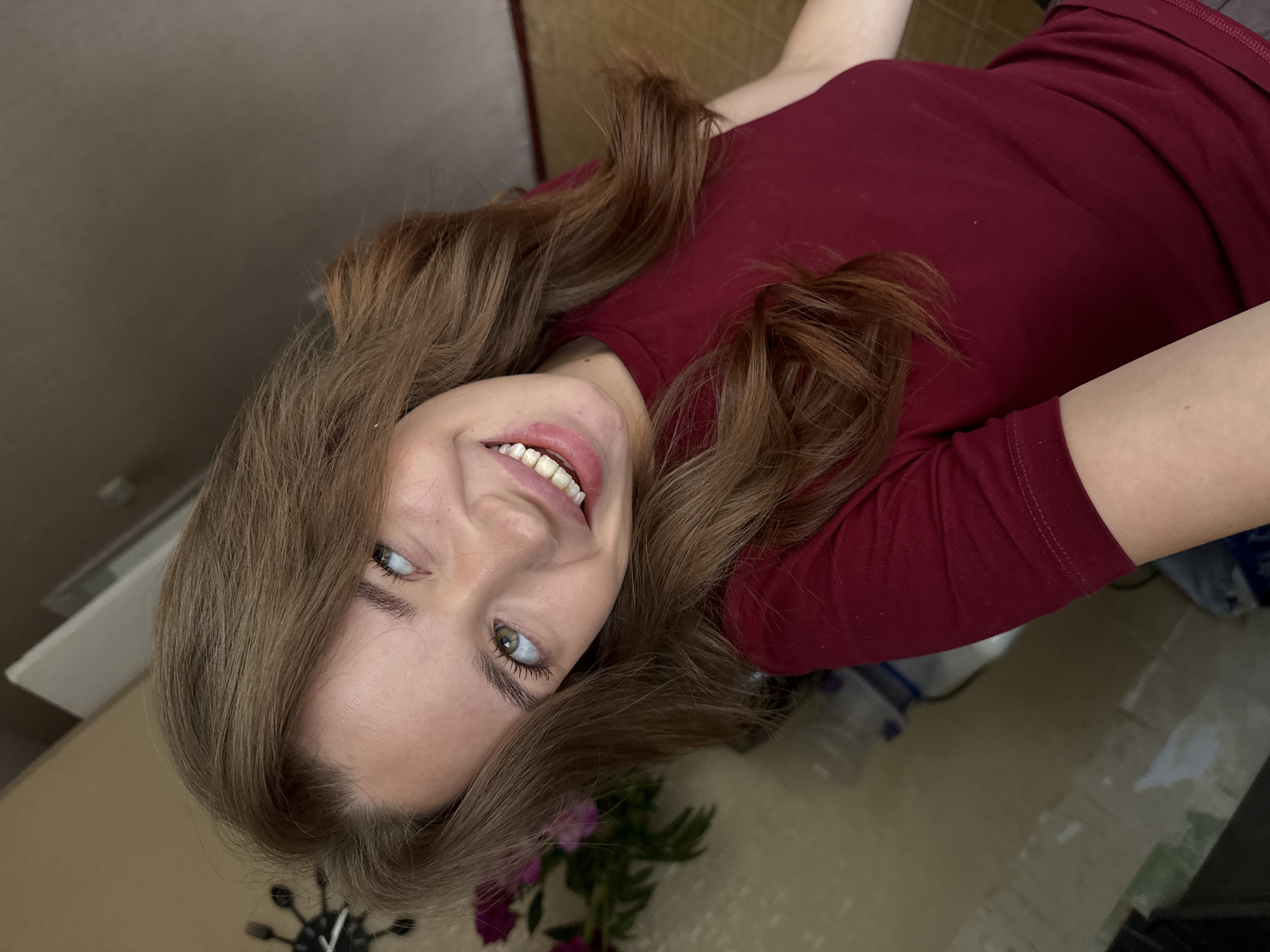 A young woman with long, wavy, light brown hair is captured in a close-up, angled selfie. She is wearing a dark red, short-sleeved t-shirt. Her head is tilted, and she is looking upwards and to her right, with a wide smile revealing her teeth. Her eyes are a light green color. Her left arm is visible, resting in the frame. In the background, on the left side, a bouquet of purple flowers is partially visible, alongside a dark, possibly metal object with protruding elements, resembling a clock or decorative piece. The background also shows a wall with textured surfaces and a hint of what appears to be flooring. The overall impression is an indoor setting, possibly a home. The image does not contain any information that would allow for its location to be identified as Chaikovsky, Russia.