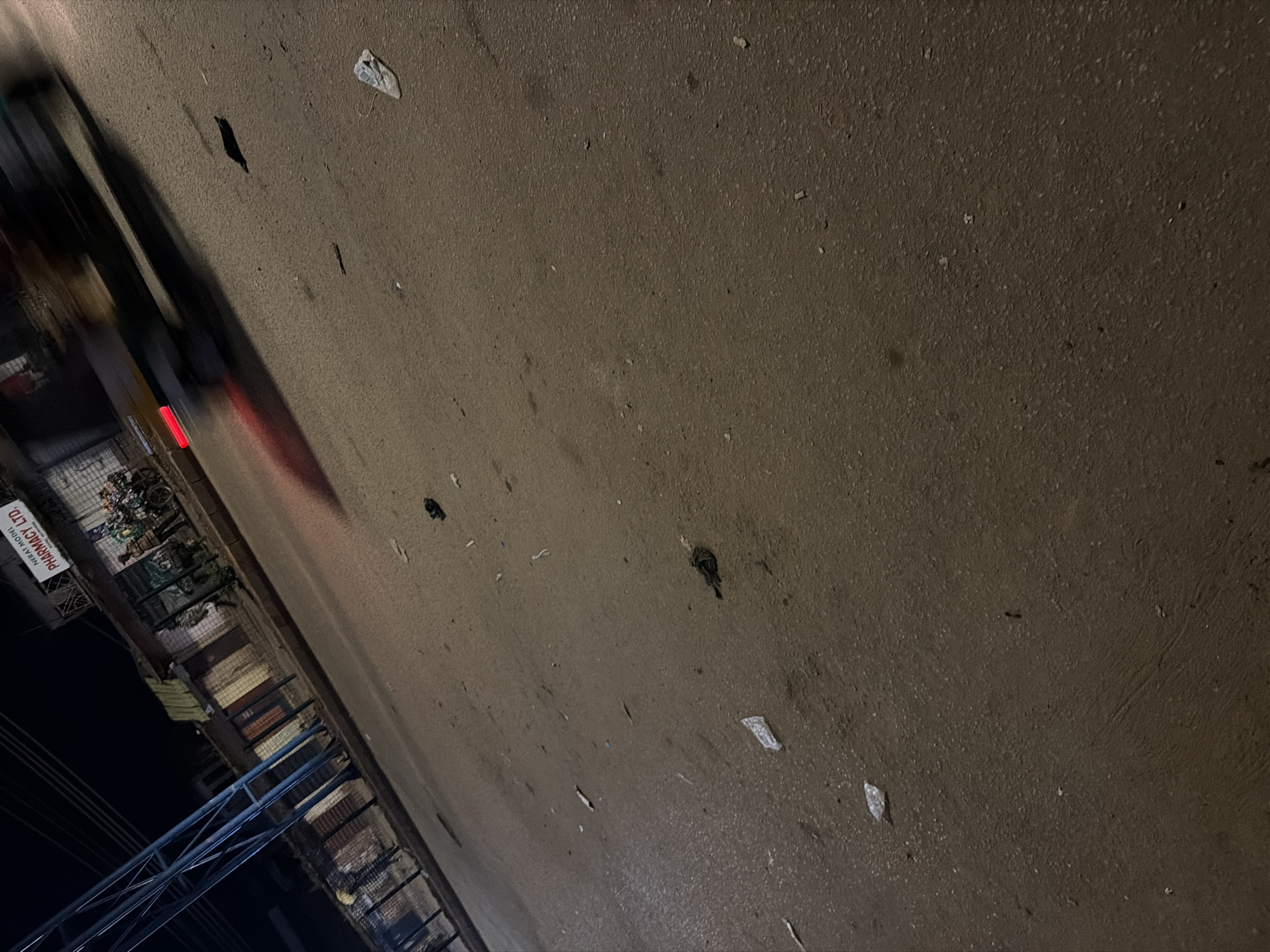 This is a low-angle, night-time photograph taken on a street in Bukuru, Nigeria. The road surface is asphalt and appears wet, reflecting the artificial lighting from streetlights and shop fronts. Debris, including plastic bags and what appear to be leaves or litter, is scattered across the road.
On the left side of the image, a tall, blue metal structure, possibly a utility pole or part of a bridge, dominates the foreground. In the background, along the side of the road, are several small shops with their shutters down or closed. One shop has a sign that reads "NIFYAT MODEL PHARMACY LTD.".
A few figures, possibly people, are visible near the shops. A motorcycle with passengers is parked in front of the pharmacy. A vehicle is captured in motion, heavily blurred due to a long exposure, its headlights and taillights visible as streaks of light. The overall scene depicts a typical urban street at night in Nigeria, with a sense of everyday activity despite the darkness and scattered litter.