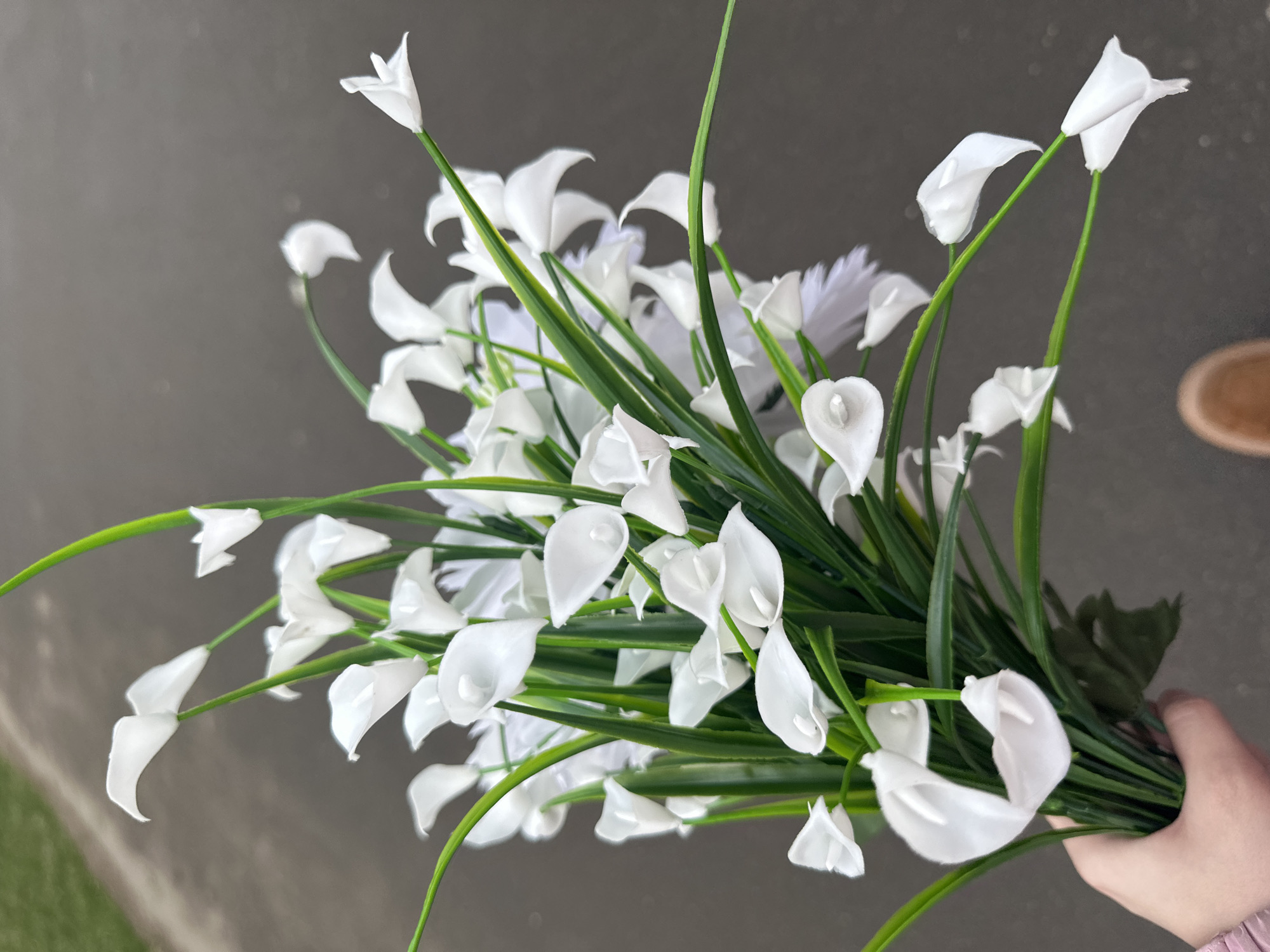 A bouquet of artificial white calla lilies with green stems is held in a person's hand. The background is a blurry mix of gray concrete and a hint of green, likely suggesting an outdoor location. A portion of a shoe is visible in the background.