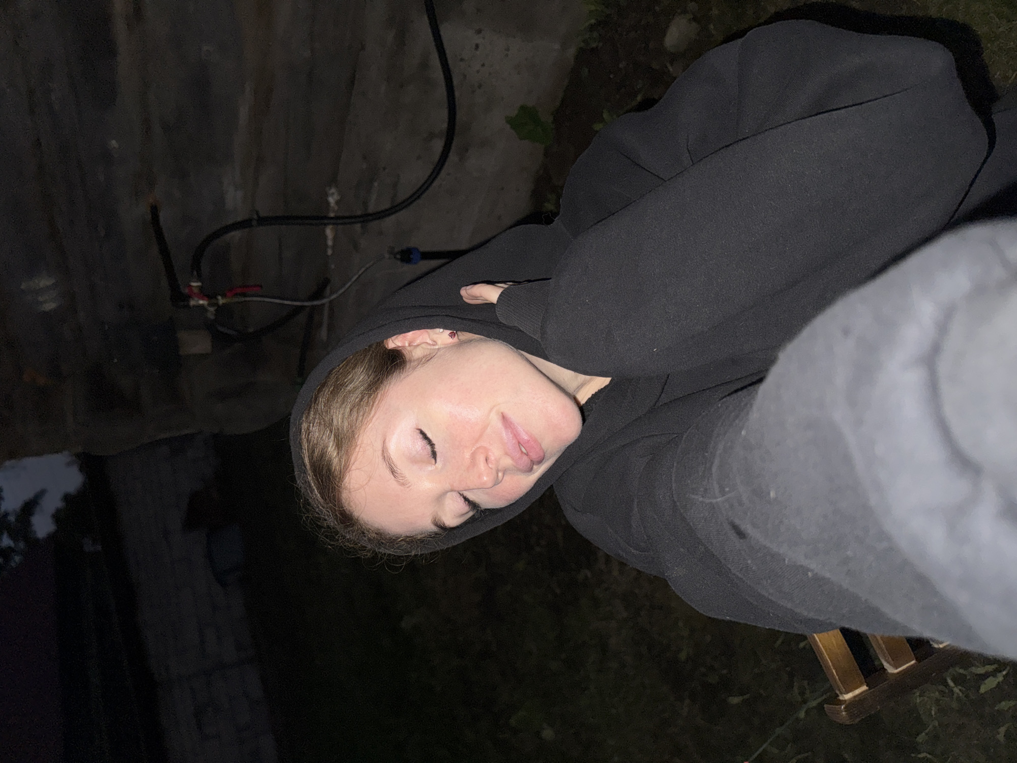 A young woman with auburn hair pulled back and wearing a black hooded sweatshirt is captured in a close-up, waist-up, low-angle selfie. Her eyes are closed, and her head is tilted back slightly, with her hands tucked into the hood of her sweatshirt, suggesting a relaxed or perhaps sleepy pose. The background is dark and somewhat out of focus, hinting at an outdoor setting at dusk or night. Visible in the background is a rough, stone-like wall with some pipes and hoses attached, and a patch of dark earth with some sparse vegetation. There is also a hint of a wooden chair on the left side of the frame. The overall lighting is dim, with the woman's face illuminated by a soft light source, possibly a flash or ambient light. The location is identified as Maksimovo, Russia.