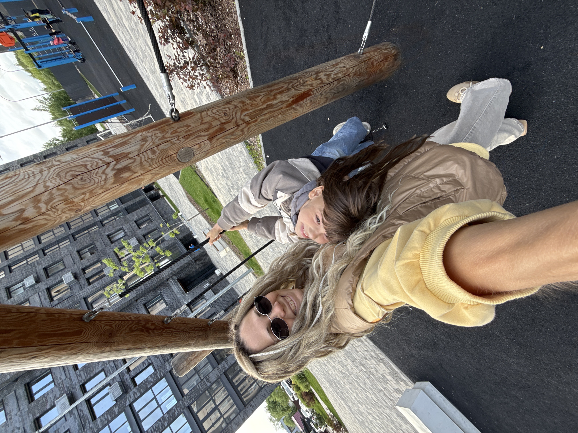 A woman and a child are on a playground, specifically on a wooden structure suspended by cables. They appear to be smiling and taking a selfie. The woman has blonde hair, sunglasses, and is wearing a yellow sweatshirt and a brown jacket. The child has brown hair and is wearing a gray and white jacket and blue jeans. The background shows a modern apartment building with numerous windows, and other parts of the playground including blue structures, along with patches of grass and pavement. The image was taken in Rusanovo, Russia.