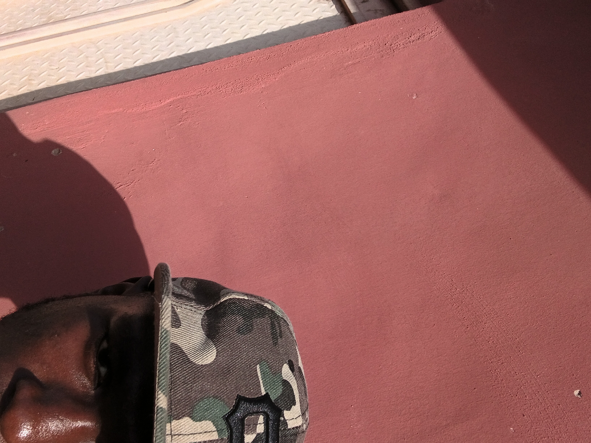 The image is a close-up, waist-up shot of a person wearing a camouflage baseball cap. The person's face is partially visible, with their right eye and part of their nose in focus. The background is a textured reddish-pink wall, with a shadow cast across it from the left. To the left of the wall, a portion of a white, diamond-patterned metal surface is visible. The overall lighting suggests an outdoor scene with direct sunlight creating strong contrasts and shadows. The context of Jalingo, Nigeria, is provided, but the image itself does not contain specific visual cues to confirm this location.