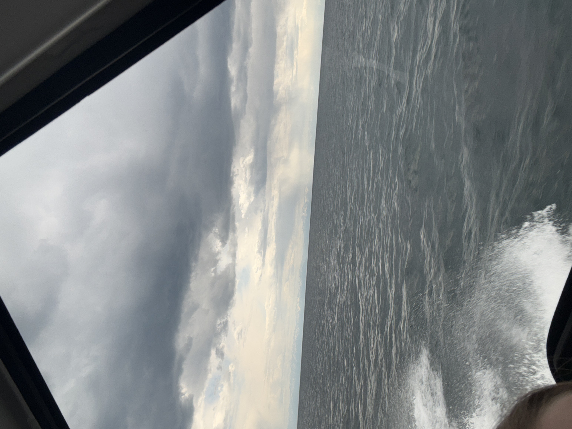 View of Lake Baikal, likely from a moving boat near Listvyanka, Russia. Visible are choppy gray water with white spray, a cloudy sky ranging from dark gray to white, and a partial view of the boat's frame.