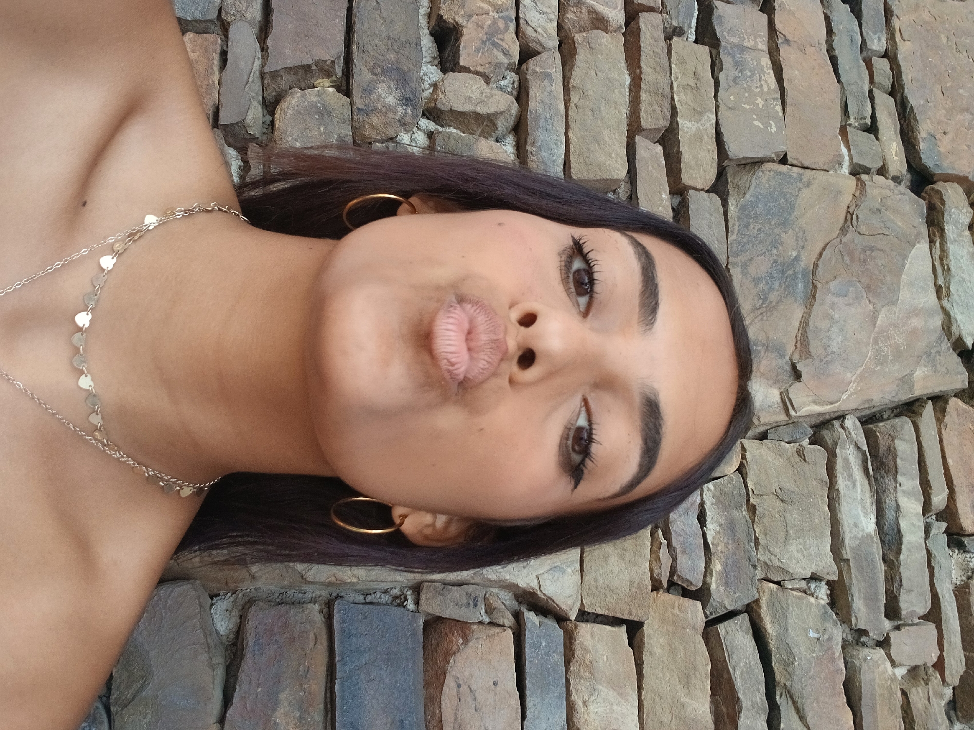 A woman with tan skin, dark brown hair, and brown eyes is seen with her lips pursed in a "duck face" expression. She wears gold hoop earrings and a delicate gold necklace with heart-shaped pendants. The background consists of a stone wall constructed of irregularly shaped, brown and gray rocks. This photograph was taken in Juan Griego, Venezuela.