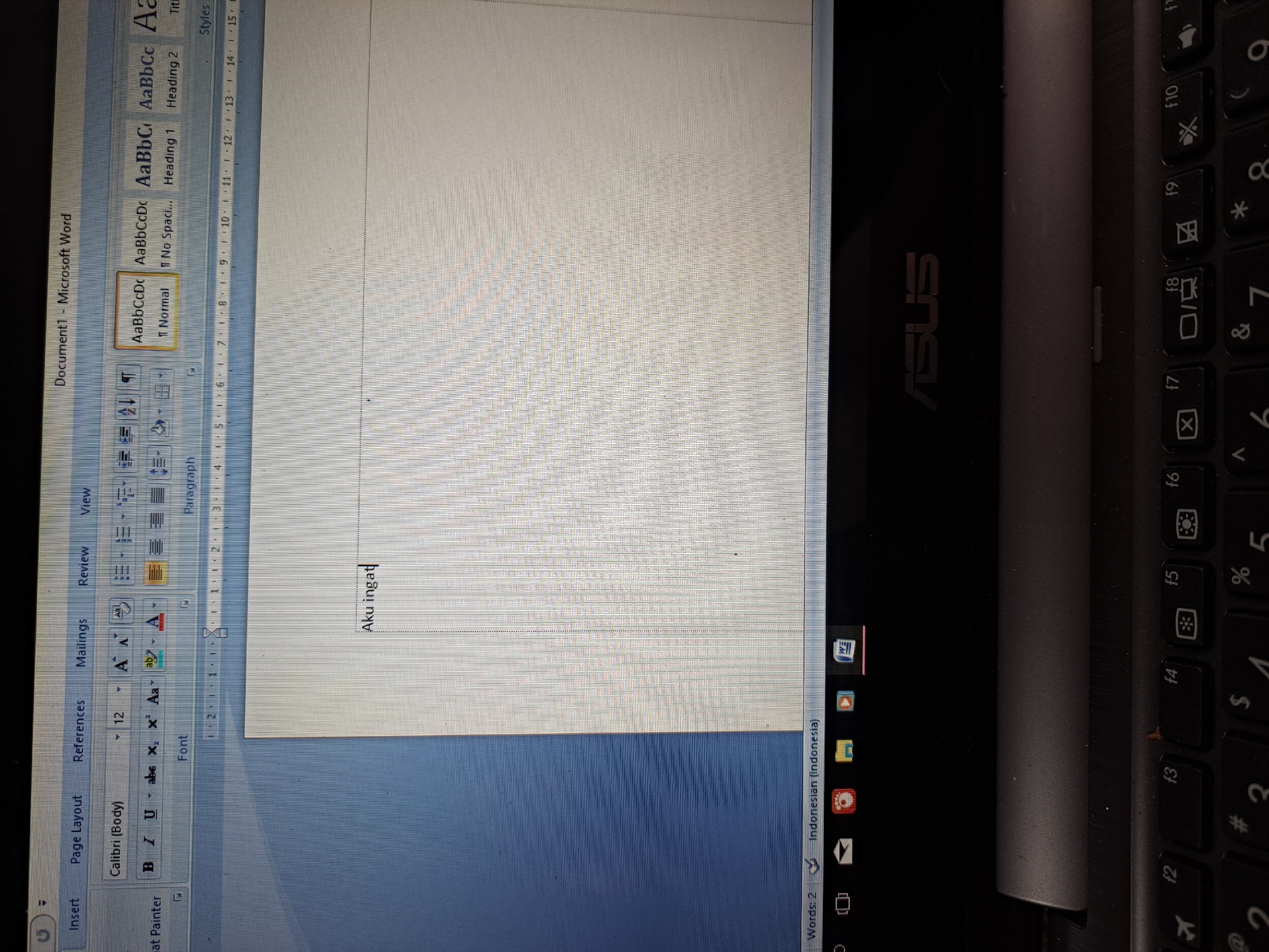 A close-up, eye-level shot shows a laptop screen displaying a Microsoft Word document. The document has the words "Aku ingat" typed on it. The top of the screen shows the Word interface with menus like "Insert," "Page Layout," "References," and "Mailings." Below these menus are formatting options and a ruler. The bottom of the screen shows a taskbar with icons and the word count and language setting. The laptop itself is dark gray, and a portion of the keyboard is visible at the bottom of the frame, with the ASUS logo also visible on the laptop's surface. The setting appears to be indoors.