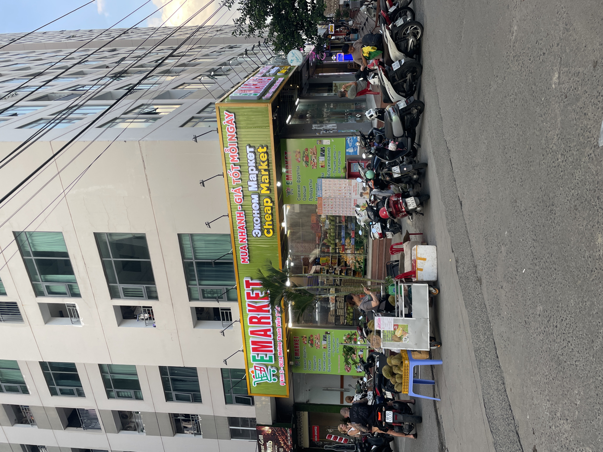 This media file captures a bustling street scene in Bac Nha Trang Ward, Vietnam, depicting a multi-story building with a market on its ground floor. The setting is an urban environment, likely a commercial street, with a concrete sidewalk and asphalt road.
The main subject is a market named "EMARKET" or "Cheap Market," prominently displayed with green and yellow signage. The sign features text in Vietnamese ("MUA NHANH - GIÁ TỐT MỖI NGÀY" - Buy Fast - Good Price Every Day), Russian ("Эконом Маркет" - Economy Market), and English ("Cheap Market"). Another smaller green sign outside lists services and products in Russian, including "Свежие фрукты" (Fresh fruits), "Овощи" (Vegetables), "Продукты" (Products), "Напитки" (Drinks), "Низкие цены" (Low prices), "Быстрое обслуживание" (Fast service), and "Лучшие цены... для туристов" (Best prices... for tourists).
Numerous motor scooters and motorcycles are parked along the sidewalk in front of the market, indicating a common mode of transport. Several people are visible: one person stands near the market entrance, possibly using a mobile phone; another is seated on a red plastic chair by the scooters. A small cart displaying durians is set up next to the market, with a person potentially attending to it. The multi-story building behind the market features many windows, some partially open, and visible power lines crisscross the upper facade. A "mur-Grill" sign is visible on a lower section of the building.
The time of day appears to be daytime, possibly late afternoon, with soft, diffused light suggesting either an overcast sky or approaching dusk. The atmosphere seems calm and typical of an urban street.