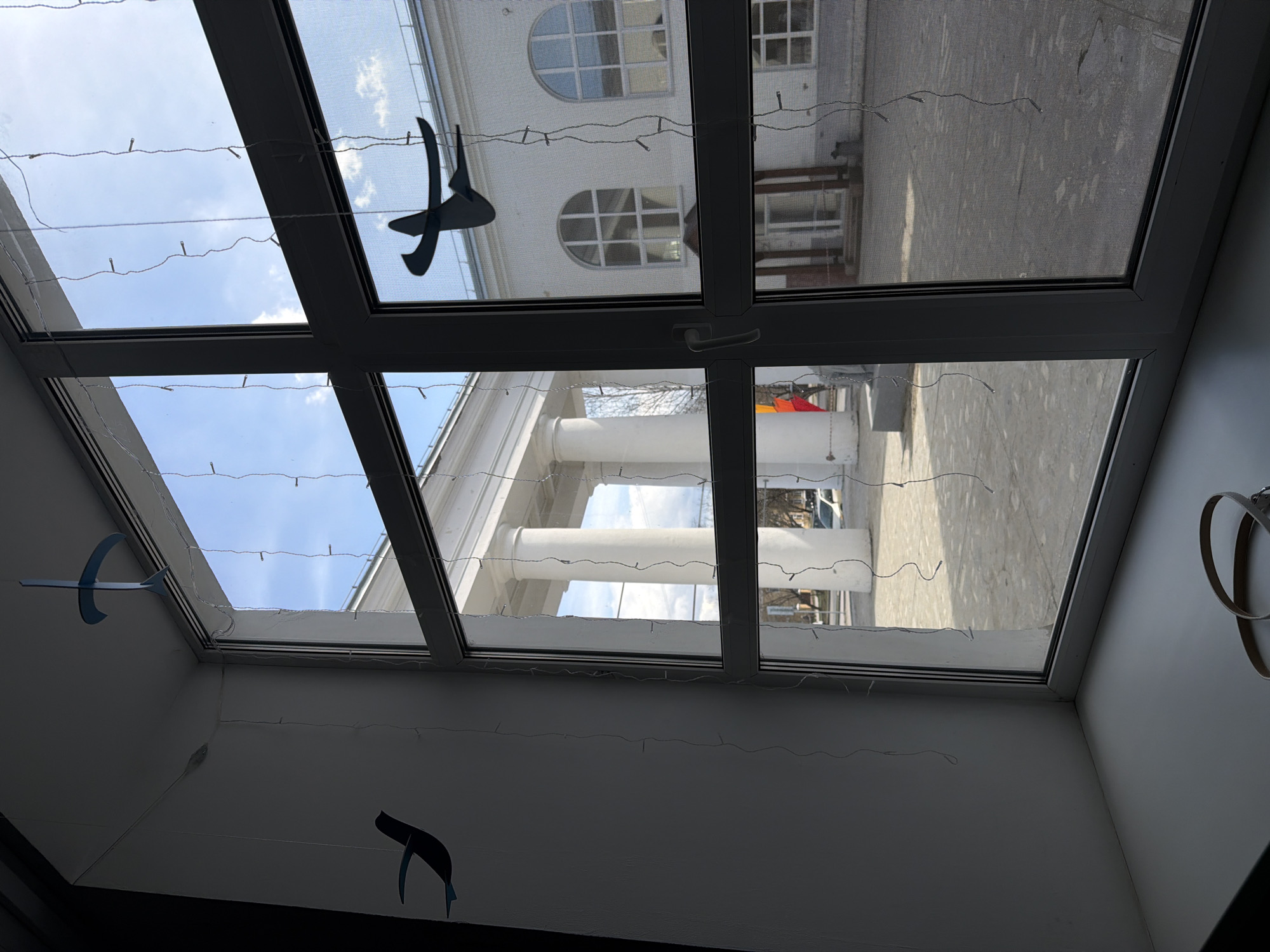 This media file presents an upward-looking view from inside a building, captured through a large, multi-paned skylight or window structure. The window frames are dark grey or black. Delicate fairy lights are strung horizontally across the interior side of the glass panes, adding a decorative touch. Several paper or cardboard cutouts, resembling abstract bird-like shapes, hang by strings from the interior ceiling or frame; some are dark, and at least one is a lighter blue.
Through the window, a bright blue sky with scattered white clouds is visible, indicating a clear, sunny daytime. The outdoor scene reveals a white, classical-style building featuring prominent white columns and arched windows with grid patterns. In front of this building, there's a paved area, possibly a courtyard or street, where a white vehicle is partially seen. Bare tree branches visible in the distance suggest the season is late winter or early spring. The interior wall visible beneath the window is white. The overall impression is of a bright day viewed from a decorated indoor space, possibly in Perm, Russia, given the location context. There is no visible text.