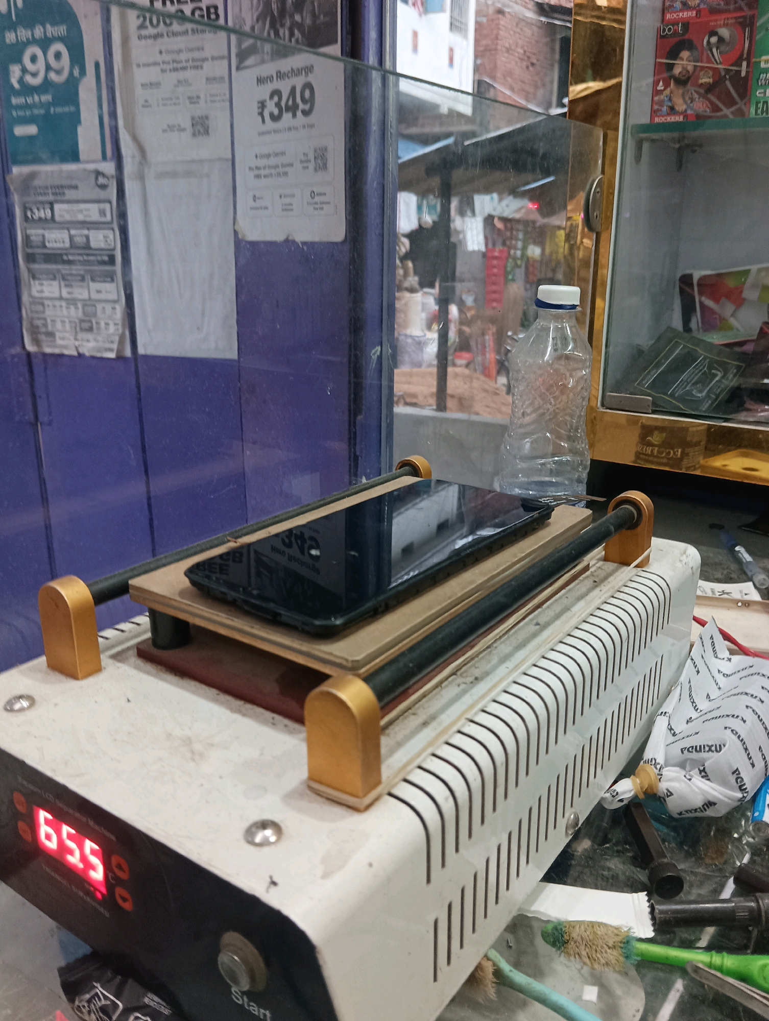 This image depicts a close-up view of a phone repair station. In the foreground, a black smartphone is positioned on a heating plate, which is part of a larger machine. The machine has a digital display showing "65.5", likely indicating temperature. The area surrounding the machine is cluttered with various tools and materials, including a plastic water bottle, a black pen, and some white paper with text. The background shows a shop interior with posters and shelves. One poster displays a price of "₹349" for a recharge. The overall environment appears to be a small business in Dakhin Parsa, India, likely a mobile repair shop, with visible signs of commercial activity and the equipment used for screen repair.