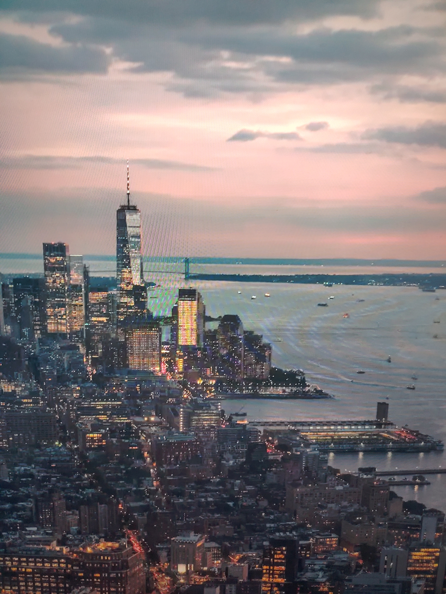 The image shows a nighttime cityscape, likely New York City, with the One World Trade Center skyscraper prominently featured. The sky above is a mix of cloudy grays and a pinkish hue, indicative of either sunrise or sunset. Below the sky, a body of water is visible, dotted with numerous boats and vessels. The city itself is illuminated with the warm glow of artificial lights from buildings and streets, creating a vibrant urban scene. In the foreground, densely packed buildings suggest a bustling metropolitan area.
