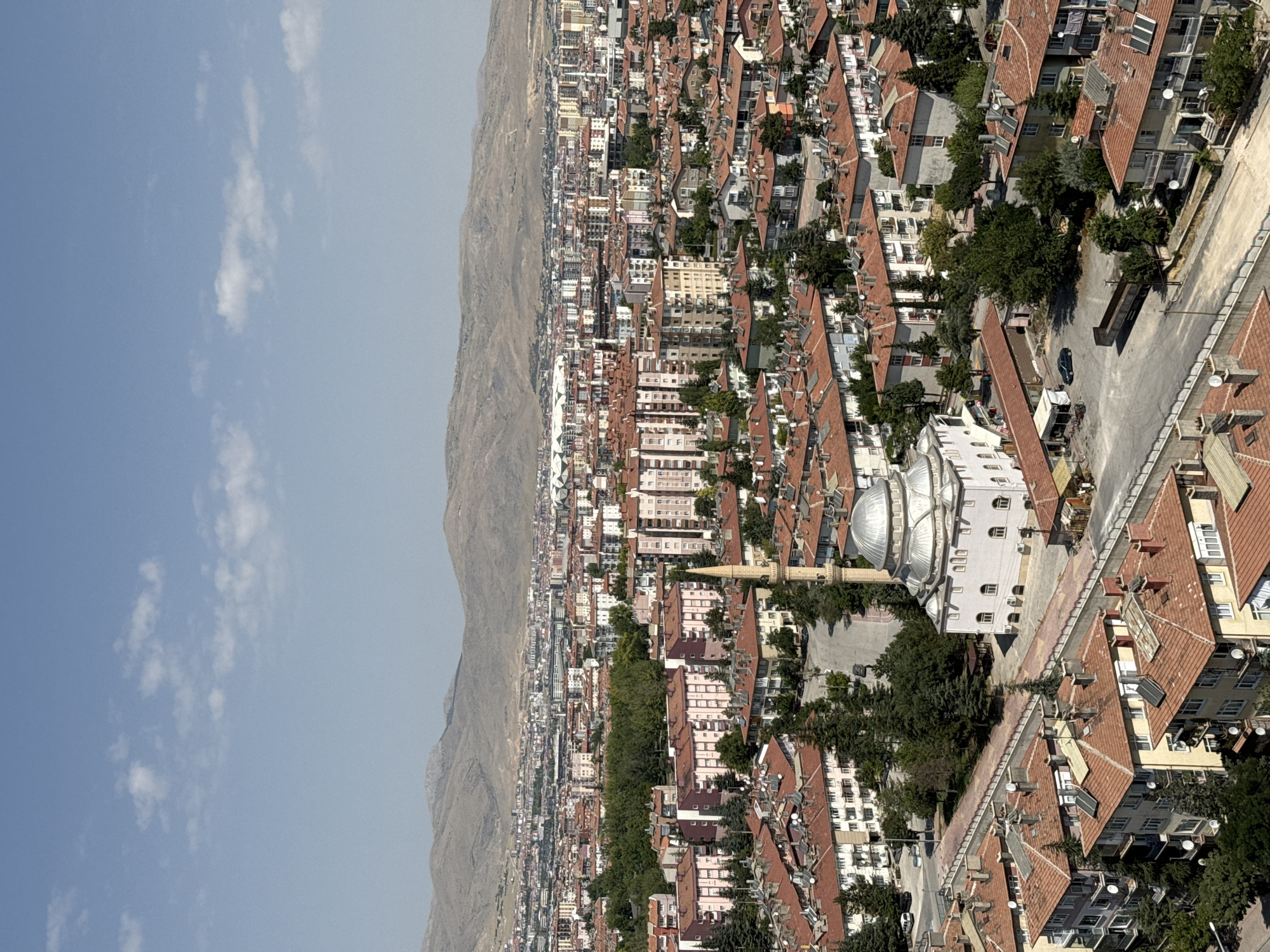 This aerial photograph captures a densely populated urban landscape in Konya, Turkey, under a clear blue sky with scattered white clouds. The foreground is dominated by residential buildings with distinctive red-tiled roofs and a variety of architectural styles, ranging from older, multi-story apartment blocks with balconies to some single-family homes. A prominent mosque with a large dome and a tall minaret is situated in the mid-ground, surrounded by trees and a paved area.
The mid-ground of the image features a significant expanse of apartment buildings, many of which are painted in light colors such as white, pink, and yellow. These buildings appear to be of recent construction, featuring multiple stories and numerous windows. A line of dense green trees creates a visual separation between this cluster of buildings and a further expanse of urban development stretching towards the horizon.
In the background, a vast cityscape unfolds, characterized by a high density of buildings of various heights and colors. A prominent feature in the far background is a large stadium with a modern, curved roof structure. Beyond the city, a range of barren, mountainous terrain forms the backdrop, its muted brown and gray tones contrasting with the vibrant urban environment. The overall scene depicts a typical residential and commercial area of a Turkish city, with elements of modern development alongside older structures.