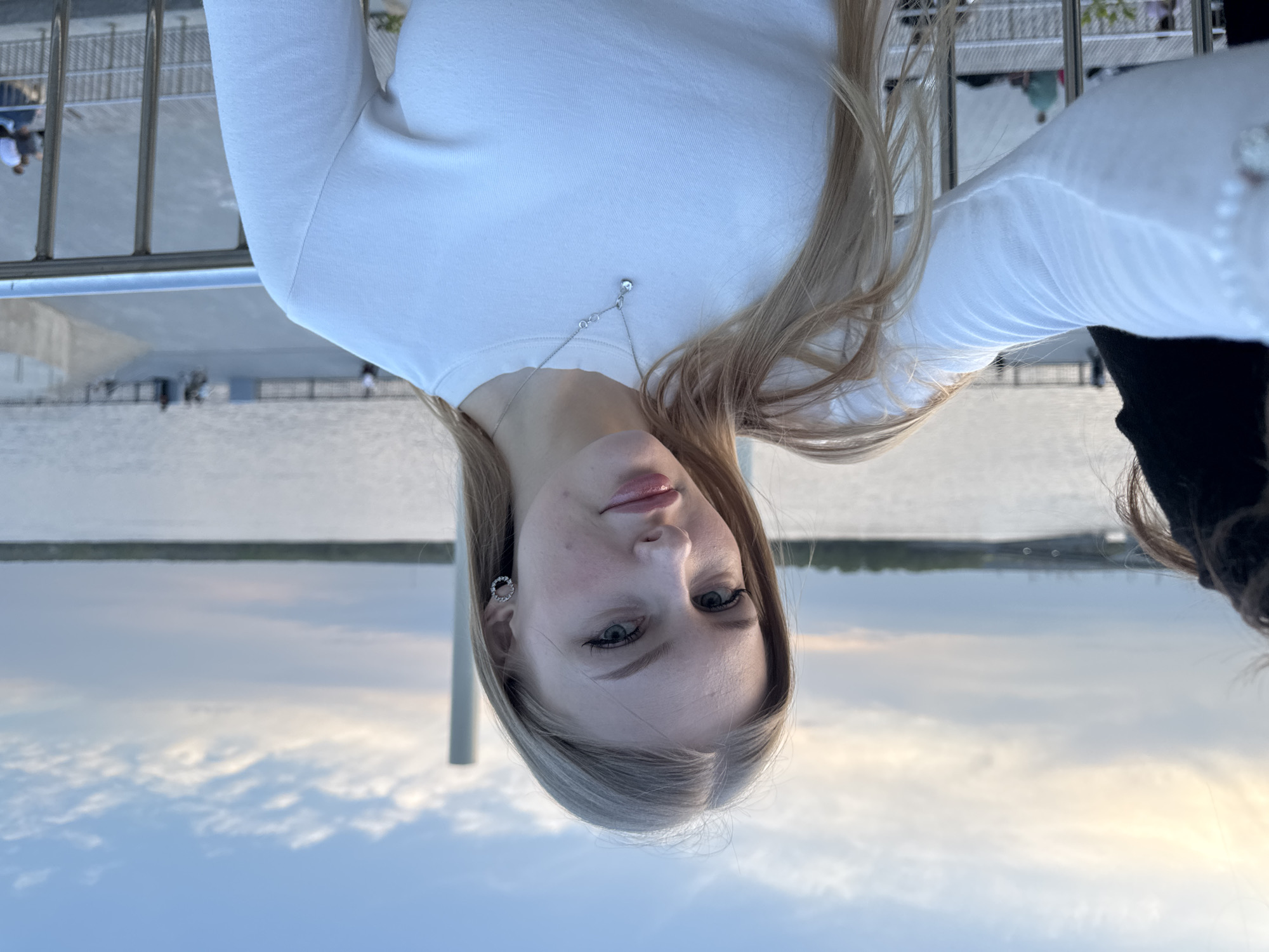 A young woman is taking a selfie outdoors in Perm, Russia. She is wearing a white long-sleeved shirt and a silver necklace and earring. Her blonde hair is long and straight. The background shows a river (likely the Kama River), a metal railing, and the city skyline under a cloudy sky.