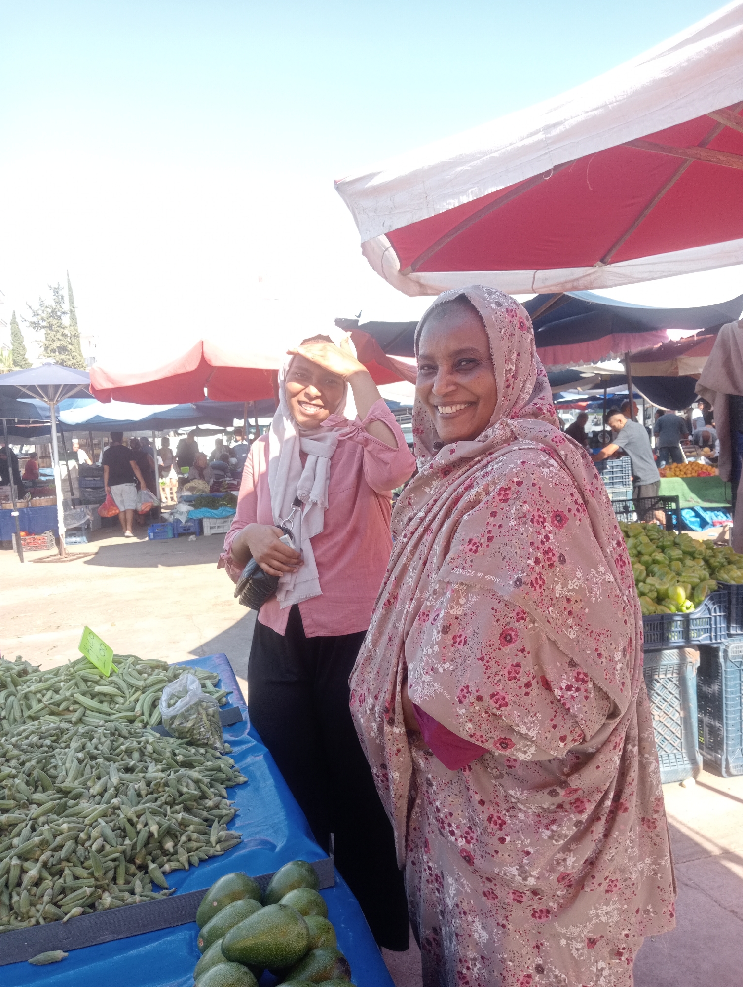 The image depicts a busy outdoor market scene in Antalya, Turkey, likely on a sunny day given the bright lighting and shadows. In the foreground, two women are prominently featured. The woman on the right is wearing a hijab and a floral-patterned shawl. She is smiling and looking towards the camera, with her right hand raised to shield her eyes from the sun. To her left, another woman, also wearing a hijab and a pink long-sleeved shirt, is visible from the chest up. She is also smiling and looking towards the camera, with her left hand holding a dark bag.
The background reveals a bustling marketplace with stalls and awnings. Various produce, including a large pile of okra and some avocados, are displayed on a blue tarp in the foreground. In the mid-ground, several people are browsing at different stalls. The stalls are covered by red and white and blue awnings. The overall impression is that of a vibrant local market day in Antalya, Turkey, filled with activity and commerce.