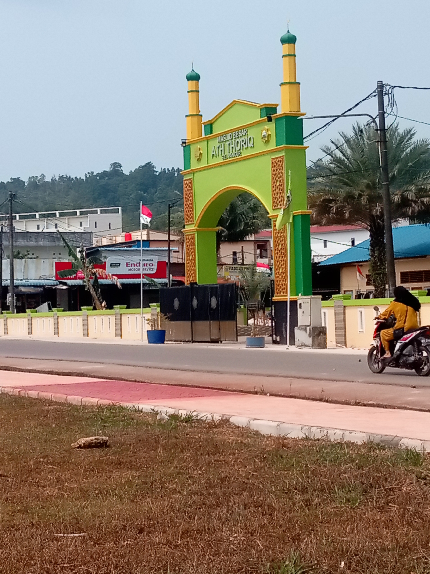 The image depicts the entrance arch of the Masjid Besar Ath Thoriq, a mosque located in Batam, Indonesia. The arch is painted in vibrant green and yellow, with decorative patterns on its pillars. A minaret, also in yellow and green, rises above the arch. Behind the mosque entrance, several buildings are visible, including one with a blue roof. To the left of the arch, an Indonesian flag is flying on a flagpole. A red and white sign for "Enduro Motor Service" is also visible on a building in the background. In the foreground, a paved road runs alongside a grassy area. A person wearing a yellow hijab and a yellow outfit is riding a motorcycle on the road, moving away from the camera. The sky is overcast. The overall scene suggests a public space in a town or city, likely during the daytime.