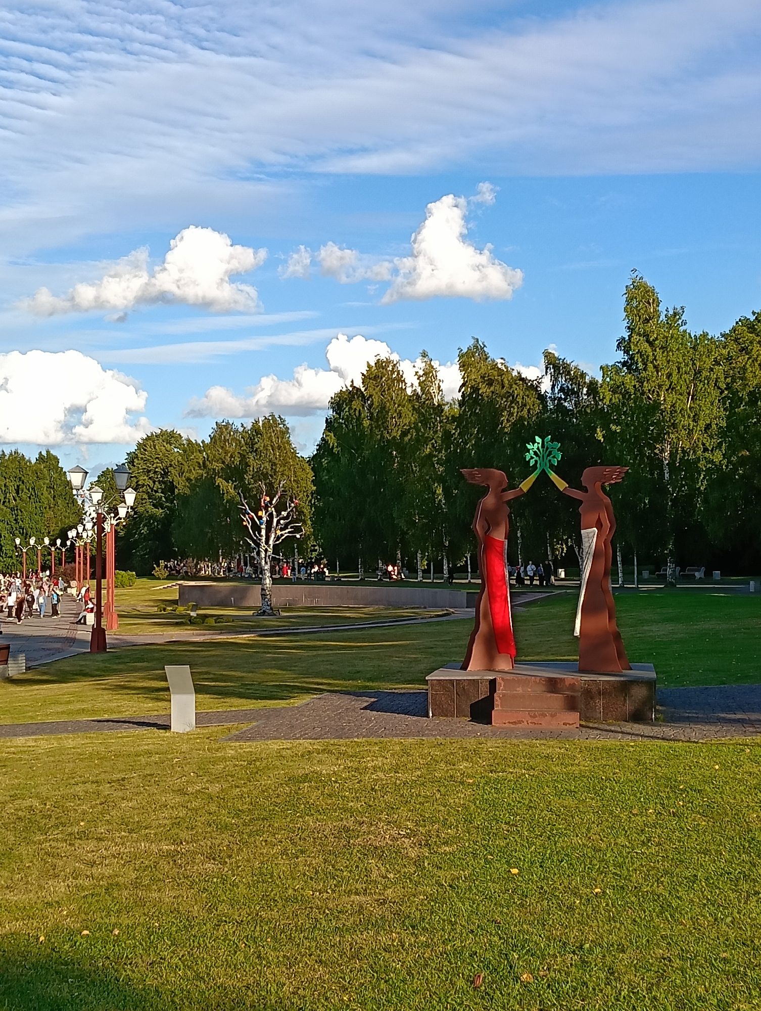 The image shows a view of a park in the city of Petrozavodsk, Russia. In the foreground, there is a statue of two figures holding a green, leafy object above their heads. The figures are made of a reddish-brown material and have draped cloths around their waists, one red and one white. They stand on a stone pedestal. To the left of the statue, a paved walkway lined with lampposts curves into the distance. People are walking on the pathway, and trees and grassy areas surround the scene. In the background, a dense line of green trees forms the backdrop, with a bright blue sky and scattered white clouds above. The sun appears to be shining, casting shadows on the grass.
