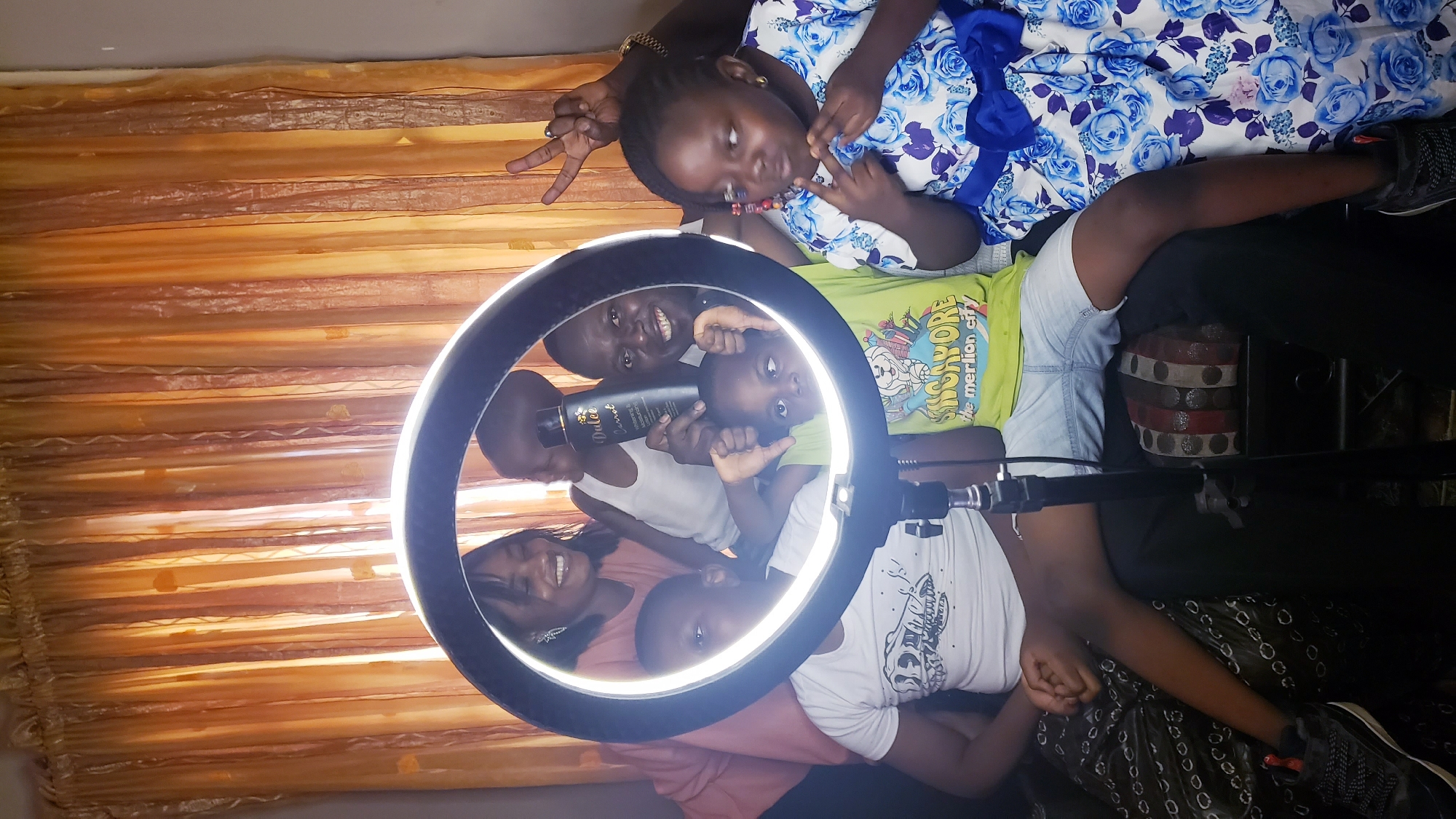 A group of people are posing for a photo under a ring light in what appears to be a home setting. The backdrop includes an orange curtain. People are holding a black bottle labeled "Dulce". A young girl in a blue floral dress is making a peace sign. Another child is wearing a green t-shirt with "Singapore" printed on it. Location details are not discernible.
