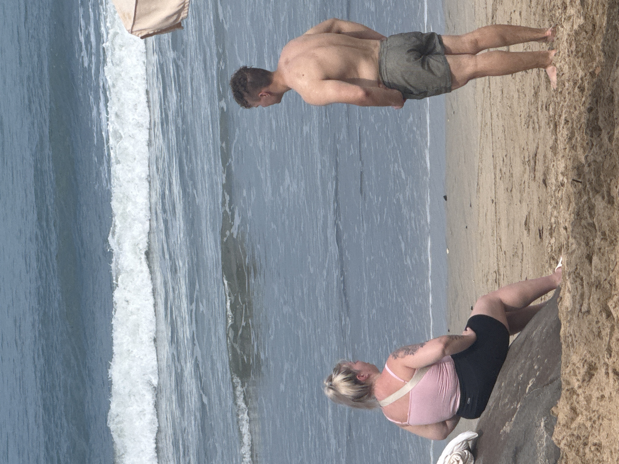 A man and a woman are standing on a sandy beach with the ocean and waves in the background. The woman is sitting on a large rock to the left of the frame, facing away from the camera and looking out at the ocean. She has blonde hair pulled back in a ponytail and is wearing a pink tank top and black shorts. The man is standing to the right of the frame, facing away from the camera and looking out at the ocean. He has short, dark, curly hair and is wearing striped swim trunks. The ocean is blue with white waves rolling in. There is a beige umbrella partially visible in the upper right corner of the image. The sand on the beach is light brown and textured. The scene appears to be a daytime shot under natural light.