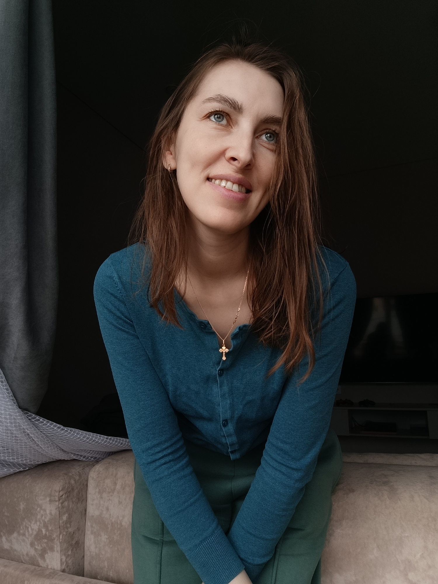 A young woman with light brown hair and blue eyes is sitting on a beige sofa. She is wearing a teal, long-sleeved, button-up shirt and green pants. She is smiling and looking upward. A gold necklace with a cross pendant is visible. Her hands are folded in her lap. Behind her, there is a dark space, possibly a window or doorway, and a partially visible curtain on the left. A television and entertainment unit are faintly visible in the background. The image was taken in the city of Chaikovsky, Russia.