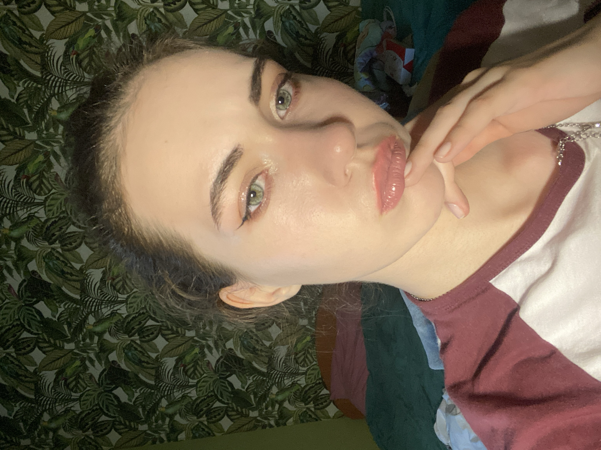 A young woman with light skin and green eyes is looking at the camera. She has dark brown hair pulled back, and is wearing makeup including eyeliner and lip gloss. Her left hand is raised to her face, touching her cheek. She is wearing a white t-shirt with dark red sleeves. The background is a green patterned wallpaper with leaves and birds. The image appears to be taken indoors, likely in a bedroom, given the wallpaper and the clothing. The lighting suggests it is daytime, but no specific time of day can be determined. No text is visible in the image.