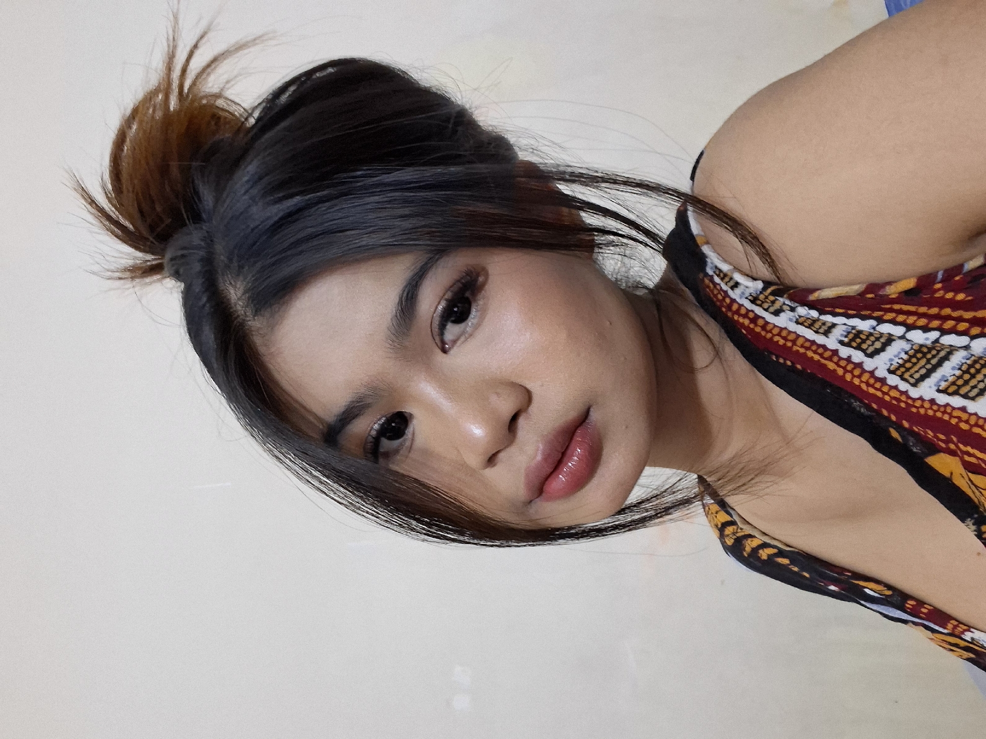 The image shows a close-up portrait of a young woman with dark hair pulled up into a messy bun. She is wearing a colorful, patterned top. Her makeup includes dark eyeliner and lipstick. The background is a plain, light-colored wall. The image is a selfie taken from a slightly upward angle. The overall setting suggests a casual indoor environment. The specific location in Kuta, Indonesia is not discernible from the image itself.