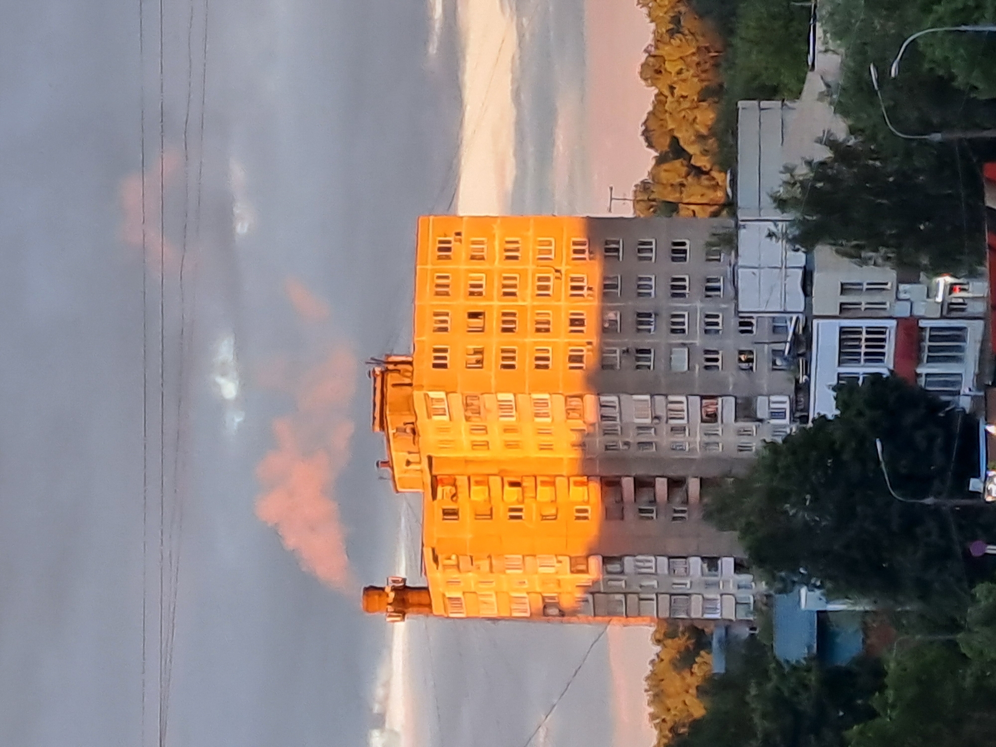 The image depicts a multi-story residential building in Elektrostal, Russia, captured during a sunset. The building is predominantly orange on one side due to direct sunlight, while the other side is gray, showing the shadow. There are numerous windows visible on both sides. A smoke stack, possibly from a nearby industrial facility, is visible. To the right of the building, there is a mix of green trees and possibly other residential structures, partly obscured by vegetation. A street light is also visible. In the background, the sky transitions from gray to orange and pink hues, indicating the sunset. Power lines are visible in the sky on the left of the image.