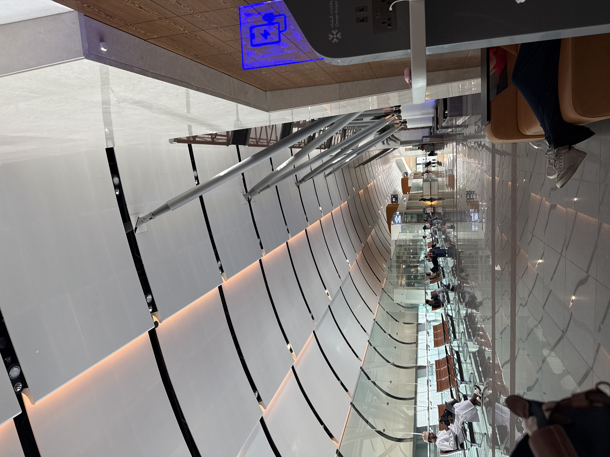 This is a photograph taken inside a modern airport terminal in Seeb Province, Oman. The image captures a wide view of the departure lounge, characterized by its high, vaulted ceiling with a segmented, wave-like design and integrated orange lighting. The floor is highly reflective, creating mirror images of the architecture and the few passengers present.
Several individuals are seated on orange, bench-style chairs arranged in rows, facing a long glass partition that overlooks an outdoor area. Some passengers are using laptops, while others are looking at their phones or resting. One man in traditional Omani attire is visible on the left, seated with a laptop. Further down the lounge, another person is seen relaxing on an orange armchair, with their legs extended.
On the right side of the image, a charging station with a prominent blue illuminated sign displaying a phone with a lightning bolt is attached to a wall panel. The charging station also has visible power outlets. The wall is finished with wood-grain paneling. The overall environment appears clean, spacious, and well-lit, suggesting a comfortable waiting area for travelers.