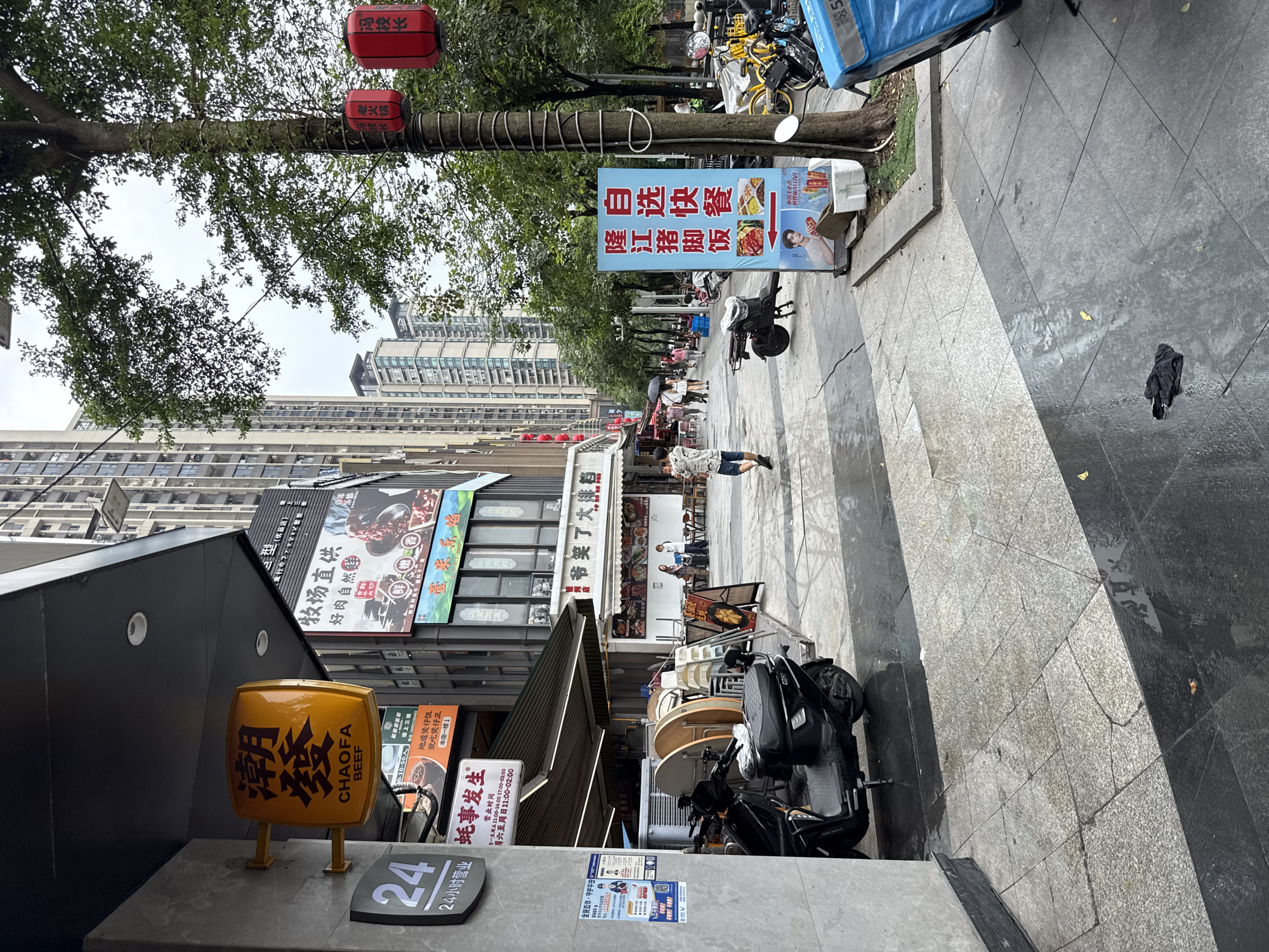 This image depicts a street scene in Haizhu District, China, likely on a slightly overcast day. The foreground is dominated by a wet, tiled sidewalk. To the left, a grey building facade with a large "24" sign is visible. An awning extends from the building, covering a row of stacked tables and chairs, with a black scooter parked in front of them. Further back, a street stretches into the distance, lined with multistory buildings and trees. A blue sign with Chinese characters and images of food is placed near a tree on the right side of the sidewalk. Several red lanterns are hung from the tree. People are seen walking and standing further down the street. A yellow delivery box is attached to a motorcycle on the far right. The overall impression is that of a commercial street, possibly a restaurant or shopping area, with signs of recent rain.