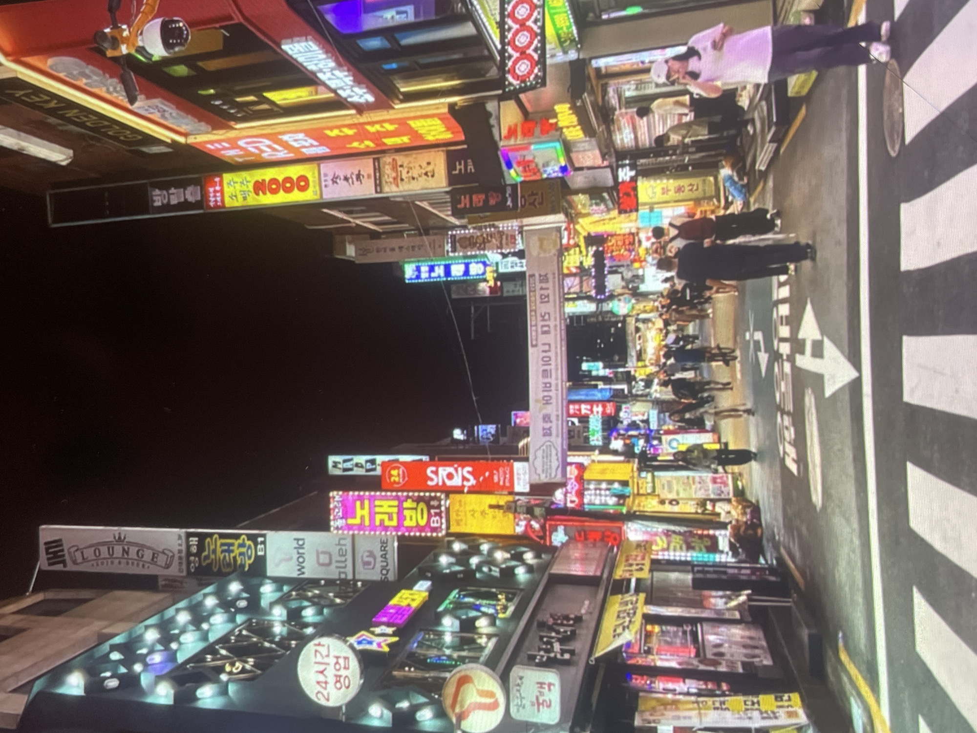 The image depicts a bustling urban street scene at night, likely in Asia, characterized by a multitude of brightly lit commercial signs in various languages, including Korean. Pedestrians are walking along a crosswalk marked with white stripes and directional arrows. The signs indicate a variety of businesses, including a lounge, a karaoke establishment, and retail shops. There is no indication in the image that this scene is in Harmanli, Bulgaria.