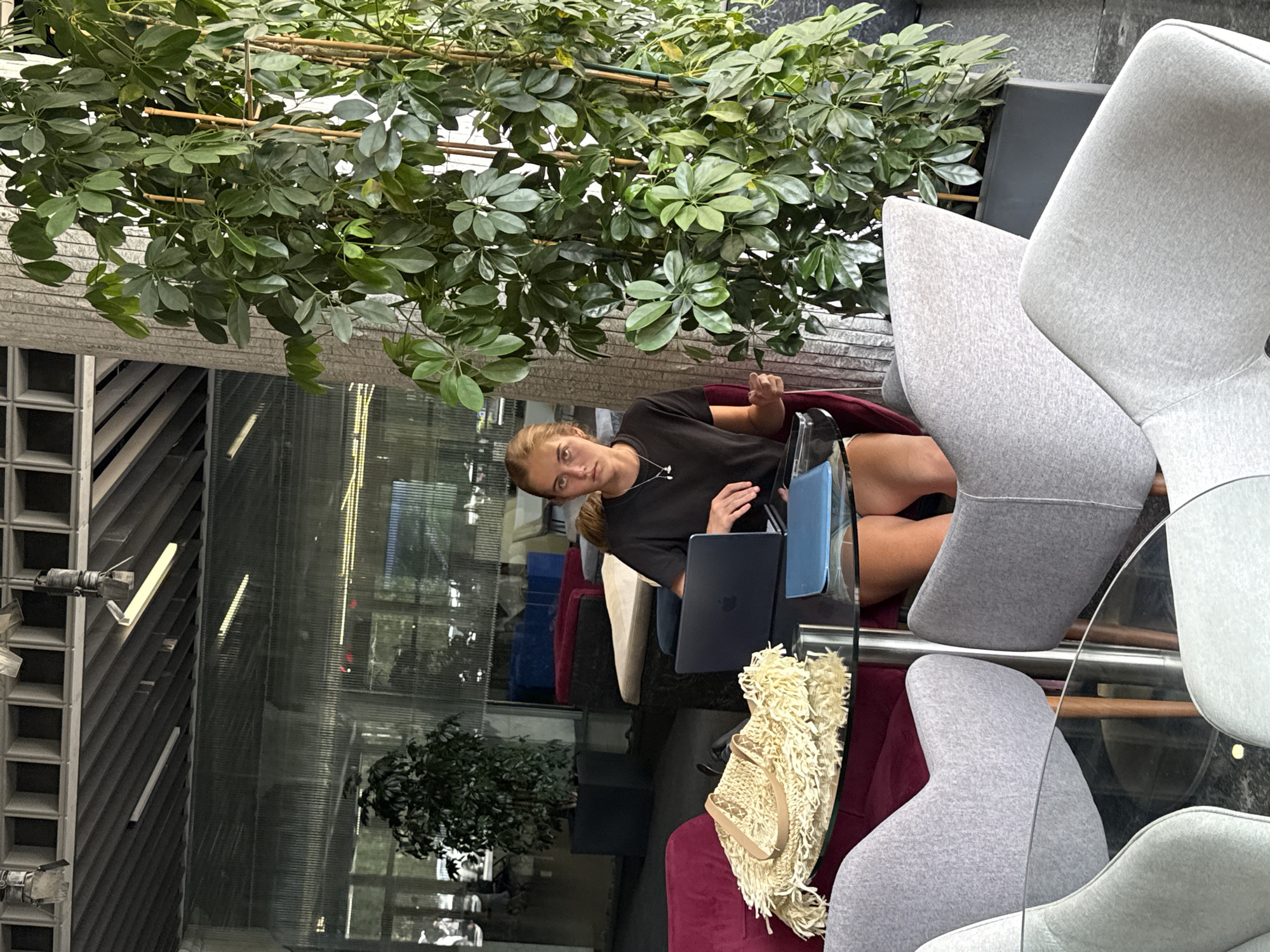A young woman with blonde hair is seated at a round glass-topped table with a laptop in front of her. She is wearing a black t-shirt and shorts. A woven straw bag with fringe detail is on the table to her left, along with a blue folder. The table is surrounded by several chairs, some upholstered in gray fabric and one in deep red. Behind her, a large green plant in a dark rectangular planter stands tall. The background shows a modern interior with large windows, a concrete column, and a ceiling with visible ductwork and lighting fixtures. The overall impression is a contemporary, well-lit indoor space. This scene is not identifiable as being located in the city of Yalta, Ukraine.