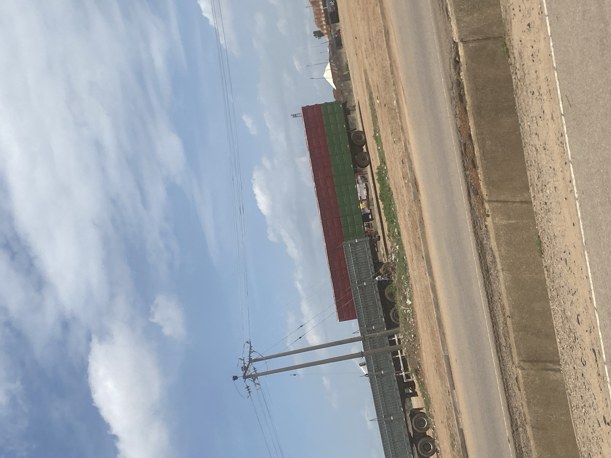 The image depicts a truck parked beside a road in Abuja, Nigeria. The truck has a multi-colored container, with sections painted red and green. An electrical pole stands nearby, with wires extending into the sky. The road surface appears to be asphalt, with a concrete barrier on the right side. In the background, there are some buildings and sparse vegetation. The sky is blue with scattered clouds.