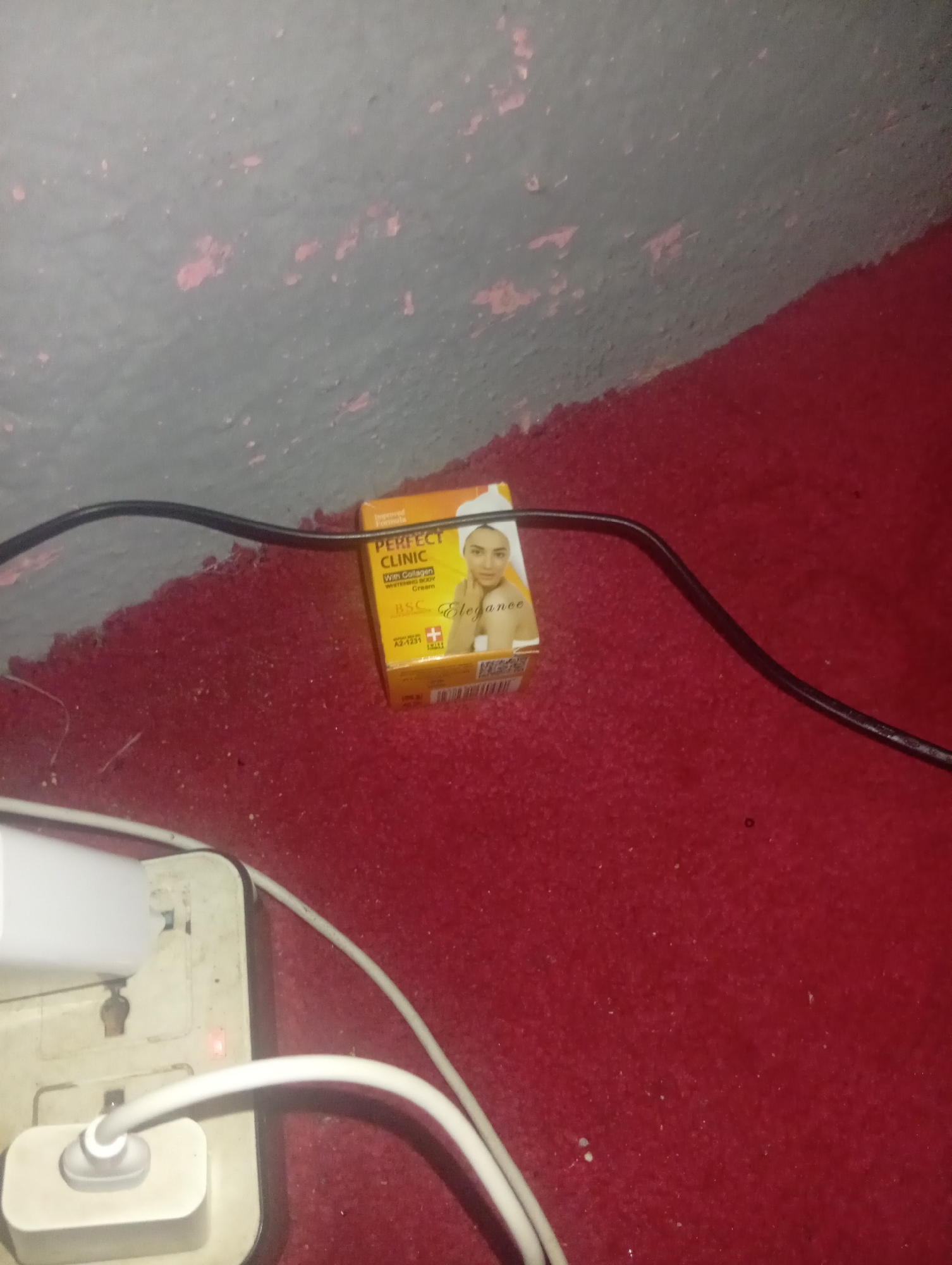 In the city of New Stadium, Nigeria, a close-up shot captures a small orange box of "PERFECT CLINIC Whitening Body Cream With Collagen" resting on a textured red surface, possibly carpet or fabric. A black cable snakes across the frame, passing near the cream box. In the lower left corner, a white electrical adapter with a red indicator light is connected to white cables, suggesting a power source. The background wall is a textured grey with peeling pink paint. The overall scene appears to be an indoor setting, possibly a domestic or storage area, with low light conditions.