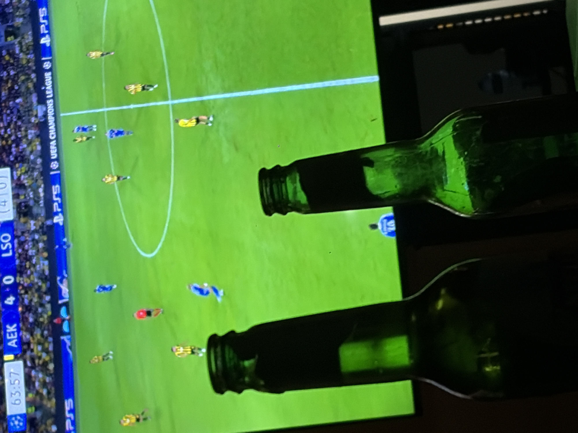 In Grigorevo, Bulgaria, two dark green glass bottles stand in the foreground, silhouetted against a television screen displaying a football match. The screen shows a live broadcast of a UEFA Champions League game between AEK and LSO. The score is 4-0 to AEK, with 63 minutes and 57 seconds elapsed in the game. Several players in blue and yellow kits are visible on the green pitch. The overall lighting suggests it is evening or a dimly lit room.