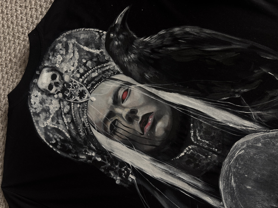 This image is a close-up photograph of an artwork painted or printed onto a black fabric, likely a t-shirt or similar garment, which is laid flat on a light-brown, textured surface.
The artwork depicts a stylized female figure with long, straight, white hair cascading down both sides. Her face is pale with a solemn expression. Her visible left eye is intensely red, and dark, tear-like streaks run down from both eyes. Her lips are slightly parted and have a faint reddish tint. She wears an elaborate, ornate grey and white headdress, possibly a kokoshnik, adorned with patterns. A small skull is positioned centrally on the headdress, above an intricate dark emblem. The figure's attire is dark with intricate, possibly silver-colored detailing visible around the neck and collarbone area.
To the female figure's right (viewer's left) stands a large, black raven. Its head is turned to the right, away from the female figure. A rounded, grey object is partially visible in the bottom left corner of the frame, underneath the female figure's left side.
Regarding the location: The image provides no visual information, such as landmarks, geographical features, or distinctive architectural elements, to identify its physical location in any city, including Krasnoyarsk, Russia.