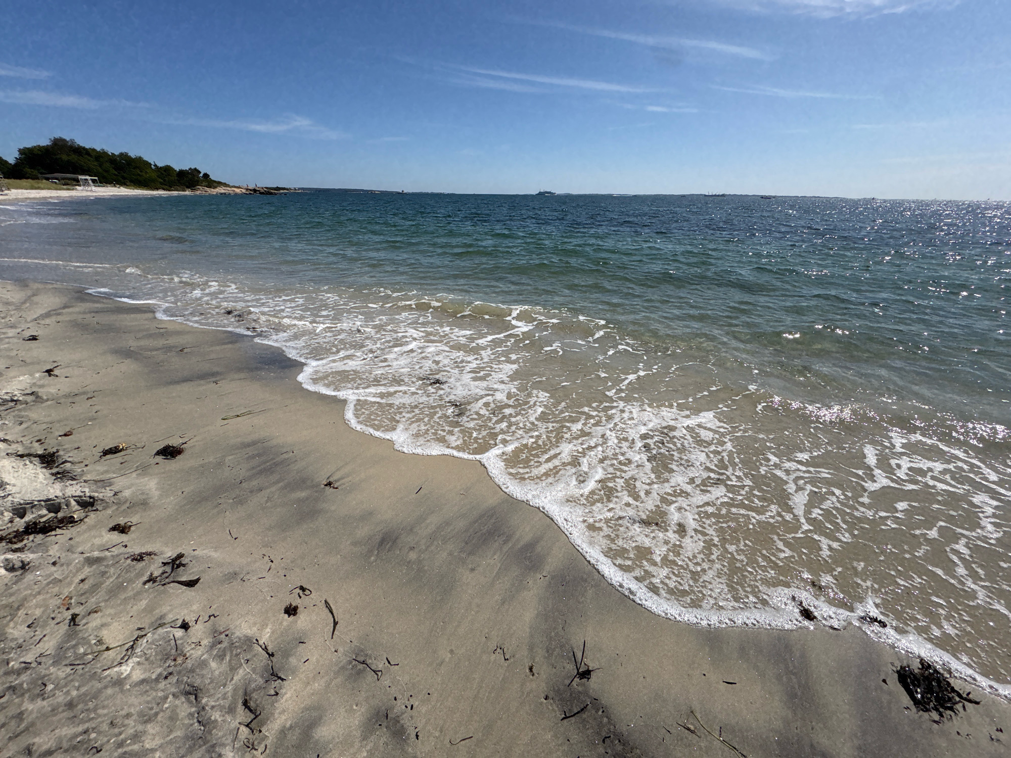 This image captures a view of a sandy beach with gentle waves lapping at the shore. The water is a deep blue with hints of green, and the sky above is a clear blue with scattered wispy clouds. The beach is composed of light-colored sand, with some dark seaweed and debris scattered across its surface. In the distance, a line of green trees and some rocky outcroppings can be seen along the coastline. The scene evokes a sense of tranquility and natural beauty. Based on visual cues such as the type of coastline, vegetation, and the presence of a lighthouse in the distance (which is a common feature in coastal areas of Waterford, Connecticut), this scene is likely located at Ocean Beach Park in Waterford, Connecticut.