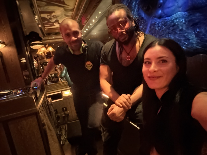 The image depicts three individuals inside the Blue Room of Bob Bob Ricard Soho, located in London, United Kingdom. The dimly lit interior features dark wood paneling, decorative warm-toned wall sconces, and a reflective ceiling with an abstract pattern. A prominent large, blue-lit, textured sculptural element resembling a gorilla head and torso dominates the right background.
In the foreground, a woman with long dark hair and fair skin, wearing a dark top, is positioned closest to the camera, smiling directly at it, suggesting she is holding the camera for a selfie. To her left and slightly behind, a man with dark dreadlocks and a beard, wearing pink-tinted sunglasses, a black v-neck top, and a red circular pendant, holds the woman's left arm with his left hand while looking towards the camera. To the far left, behind a dark wooden DJ booth, a bald man with a light skin tone, wearing a black t-shirt with a yellow emblem and headphones around his neck, is operating a turntable with his right hand. He also looks towards the camera.