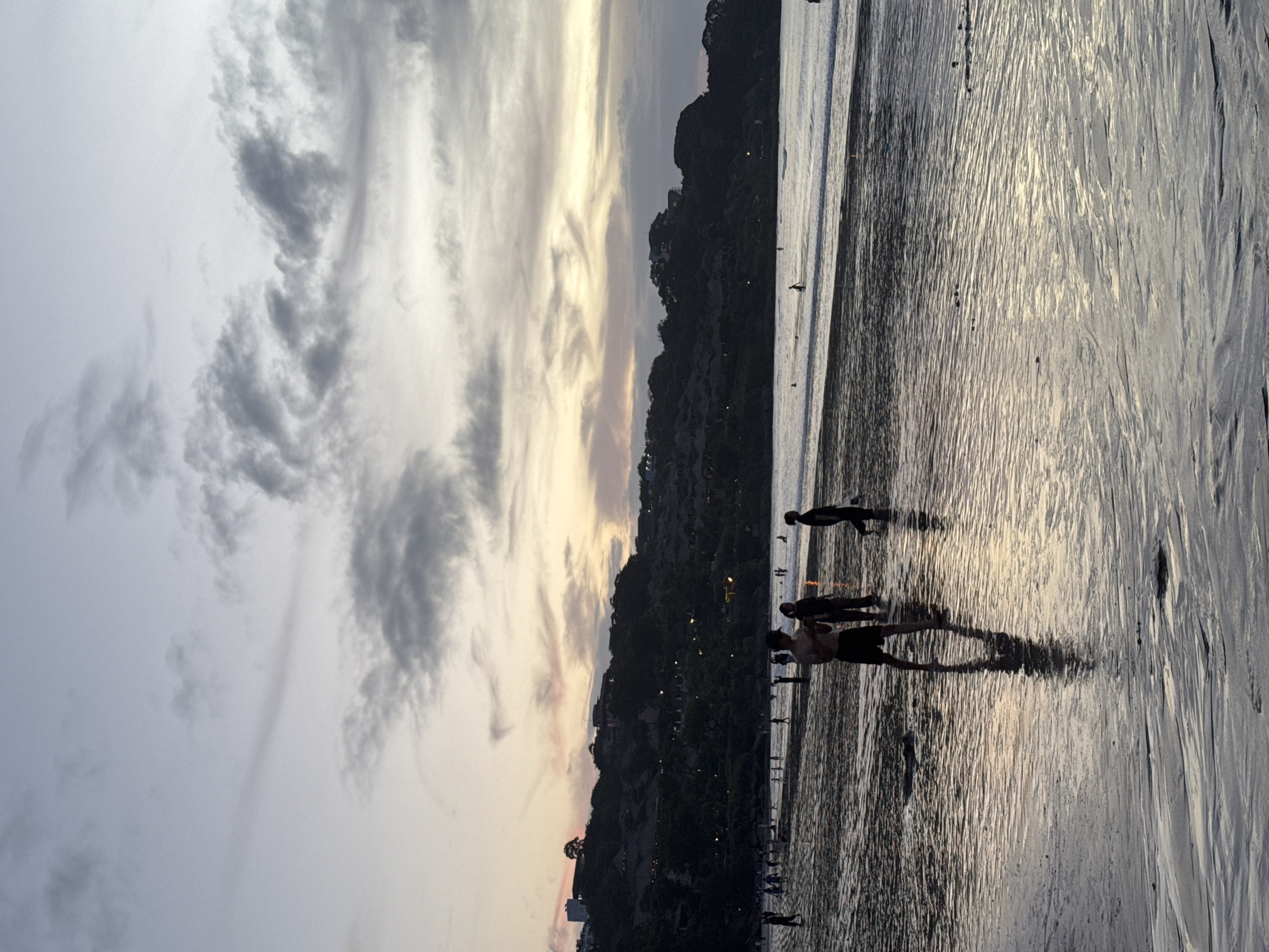 The image shows a beach scene with people silhouetted against the water and sunset sky. The sky is filled with grey and white clouds, with streaks of yellow and orange light from the setting sun. A dark, tree-covered hill or small mountain sits along the shoreline. Figures, possibly walking or playing, are visible in the water along the beach. The sand appears wet and reflective, mirroring the colors of the sky. Lights from a city on the hill are visible. The specific location within an unknown city is indeterminable from the visual information.