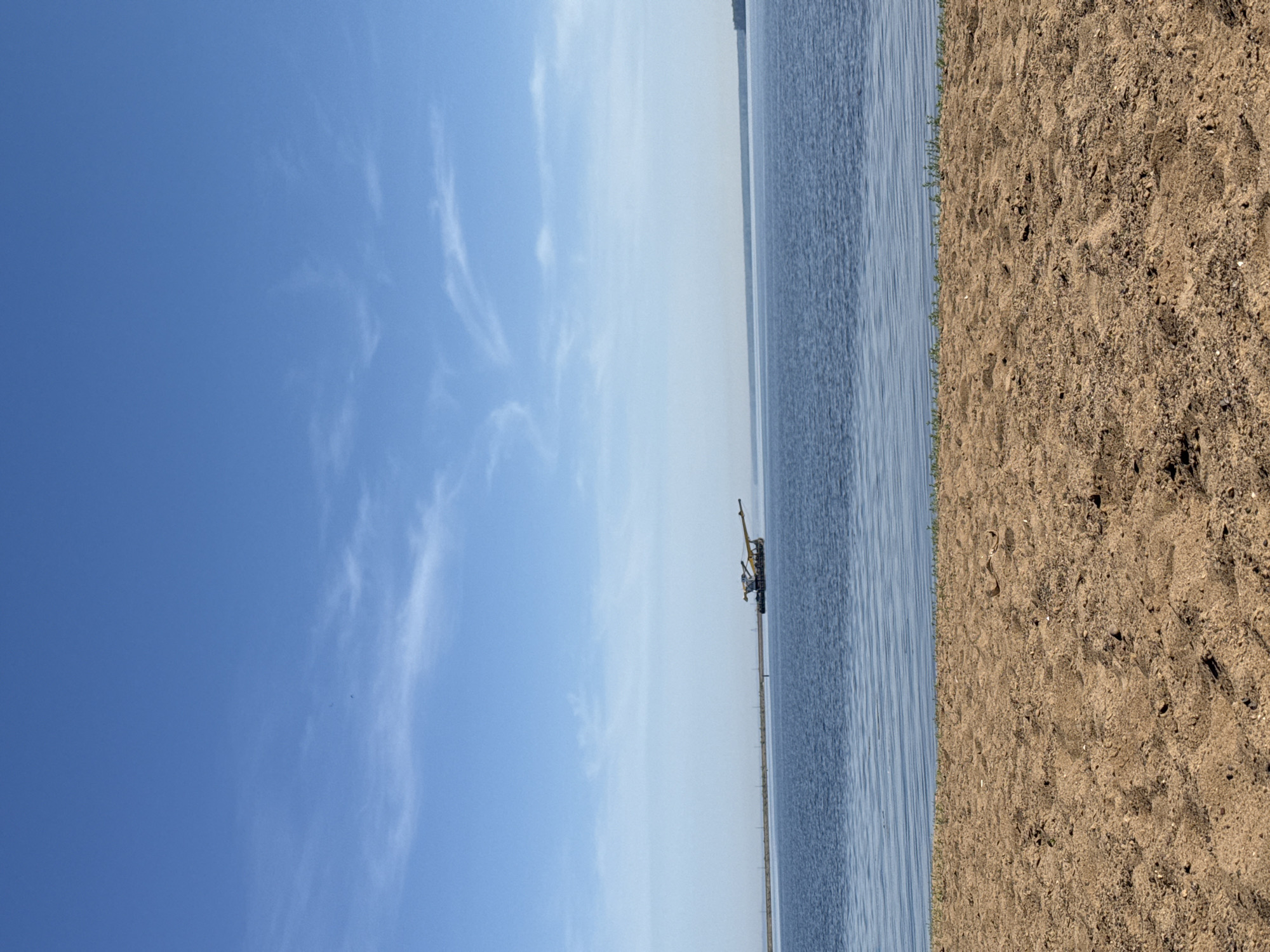 A landscape-oriented photograph shows a scene at a beach or shoreline with water, sand, and sky. The image is oriented vertically, with the blue sky occupying the upper left portion and the sandy beach visible on the right side. A stretch of water sits between the sky and sand. A boat is visible, with a crane-like structure on it. The sky is clear with some clouds. The location is likely in Chaikovsky, Russia, given the prompt's instructions. No people are evident in the scene.