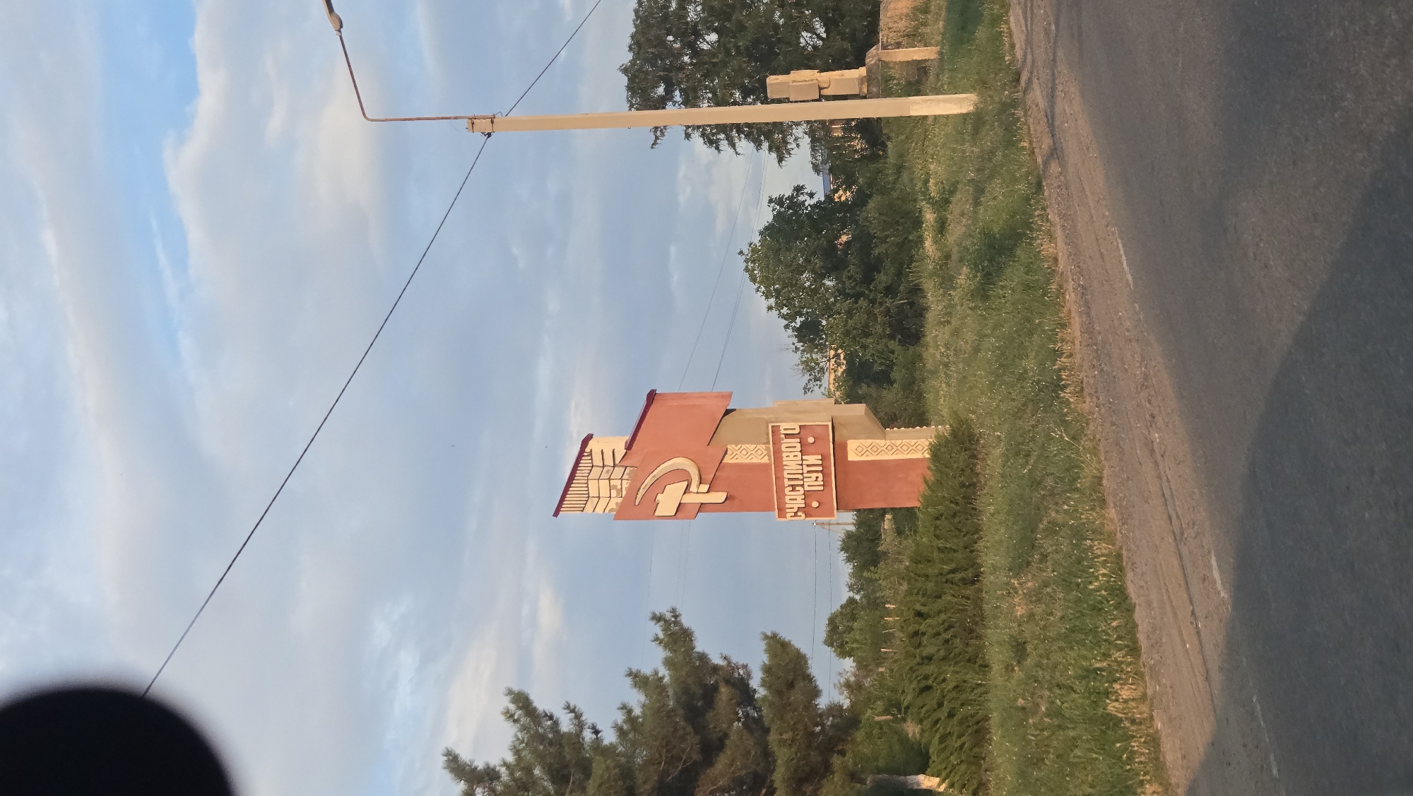 The image depicts a signpost in the city of Suklieia, Moldova, on a cloudy day. The signpost is red and beige, with a hammer and sickle symbol at the top, indicating a historical or Soviet-era influence. Below the symbol, the words "СЧАСТЛИВОГО ПУТИ" are written in white Cyrillic letters, which translates to "Happy Journey." The signpost is situated on the side of a road, with a grassy area and trees in the background. A utility pole with a street light stands to the right of the signpost. The overall scene is rural or semi-urban, characterized by natural elements and roadside infrastructure.