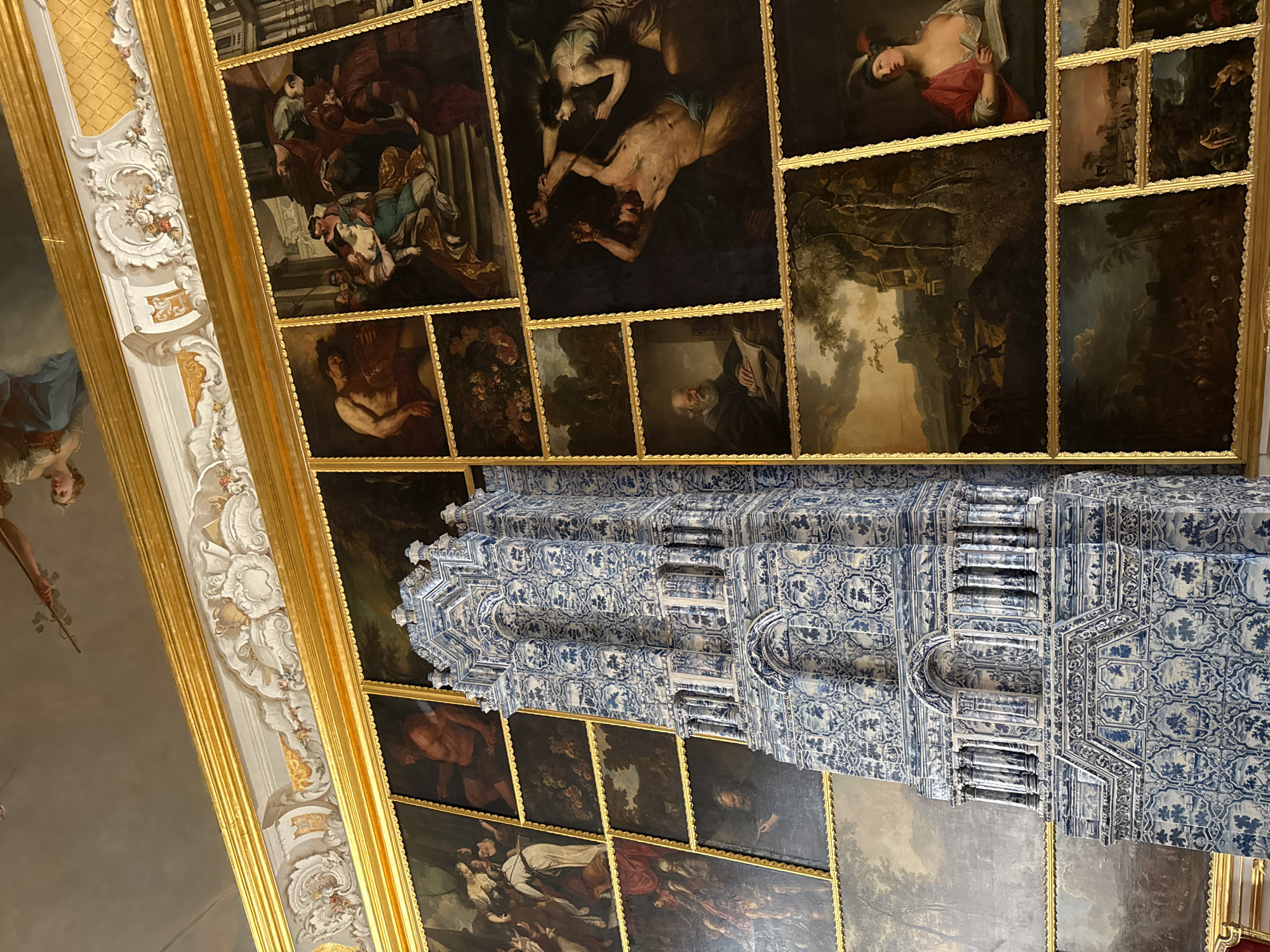 This image captures an interior view of the Catherine Palace in Pushkin, Russia, specifically showcasing a section of its opulent decor. Dominating the foreground is a tall, ornately tiled stove, predominantly blue and white in pattern, which appears to be made of majolica. This stove is positioned in front of a wall adorned with a grid of framed paintings.
The paintings themselves are varied in subject matter and composition. Several depict mythological or biblical scenes with figures in classical attire, rendered in dramatic and expressive styles. One prominent painting shows a muscular male figure bound and being tormented by another figure. Other paintings appear to be landscapes, some with pastoral elements and figures, and others are portraits of individuals, including an older man with a beard reading a book and a woman in historical dress.
The paintings are framed with elaborate gold moldings, and the entire arrangement is set against a backdrop that includes a decorative cornice and a glimpse of a frescoed ceiling with figures in the sky. The lighting in the room suggests an indoor setting, likely with ambient light enhancing the richness of the gold frames and the intricate details of the tiles. The overall impression is one of grandeur and historical richness, characteristic of a royal palace.