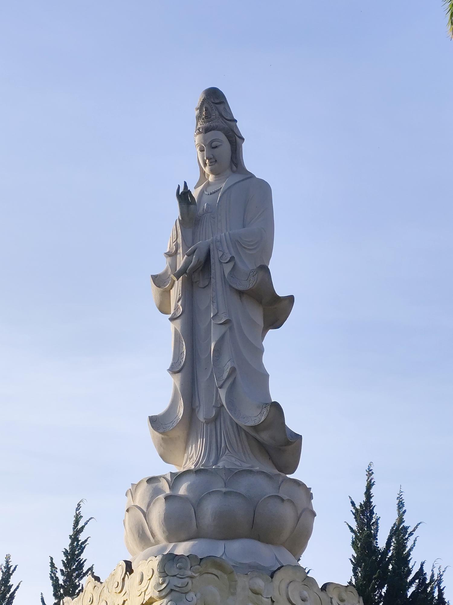 A tall, light-colored statue of a deity, likely Quan Yin, stands against a bright blue sky in Cam Ly, Đà Lạt, Vietnam. The statue is positioned on a base shaped like a lotus flower, which sits atop a decorative platform. Green, coniferous trees are visible at the statue's base on either side.