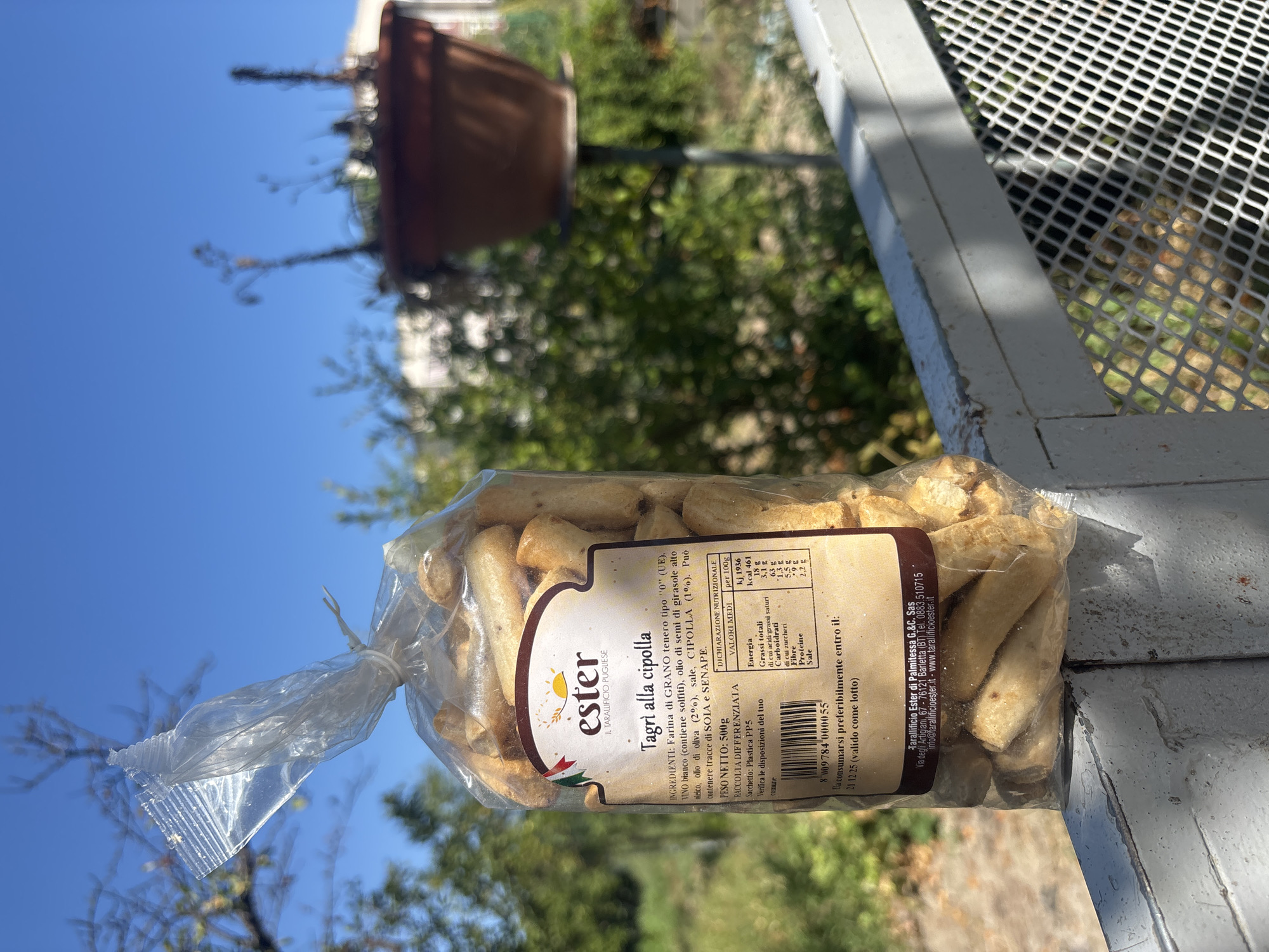 A plastic bag of "Tagli alla cipolla" taralli, a type of Italian cracker flavored with onion, is displayed outdoors, likely on a balcony railing. The bag is sealed with a white plastic tie. The label on the bag provides nutritional information and ingredient details. In the background, out-of-focus trees and a terracotta pot containing dried plants are visible against a clear blue sky. The scene appears to be located in Campomarino, Italy, as indicated by the brand name "Tarallificio Ester di Palmitessa G.&C. Sas" on the label, which lists an address in Barletta (BT), a city in the Puglia region of Italy, which is not far from Campomarino. The railing suggests a residential setting. There are no people visible in the image.