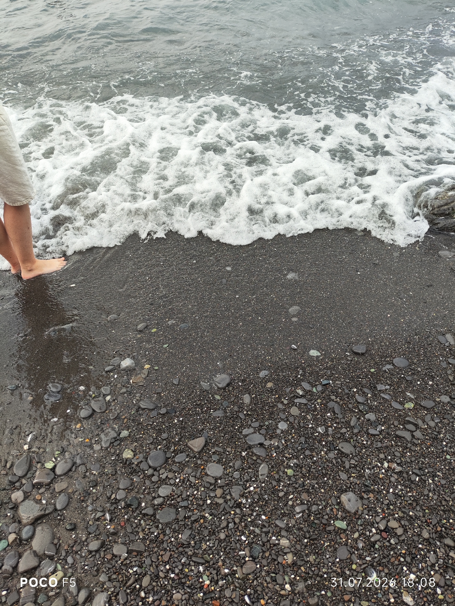 The image shows a close-up view of a pebble beach in Camogli, Italy, with the Ligurian Sea in the background. The tide is coming in, with a wave of white foam washing over the dark, wet pebbles and the feet of a person standing at the water's edge. The person is wearing light-colored shorts. The date and time stamp "31.07.2026 18:08" and "POCO F5" are visible in the lower corners of the image. The scene depicts a moment of interaction between a person and the sea on a rocky coastline.