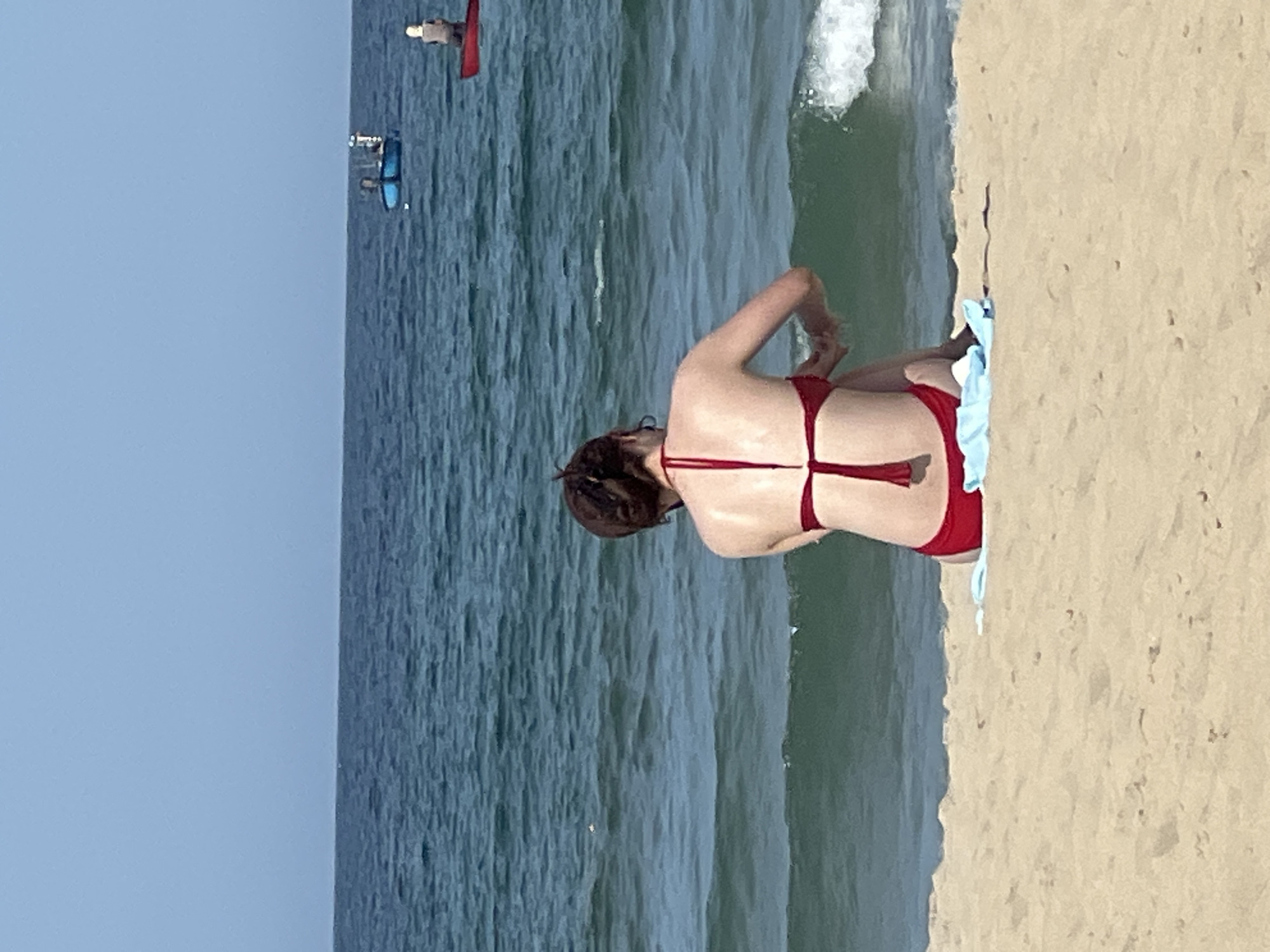 A woman wearing a red bikini sits on a sandy beach, facing away from the camera towards the ocean. She is seated on a light blue towel. Her skin is glistening, likely from sunscreen or water. The ocean is calm with gentle waves breaking near the shore. In the distance, a small blue boat with people on it and a red kayak with a person in it are visible on the water. The sky is clear and blue. The scene appears to be on a beach in Đà Nẵng, Vietnam, given the context provided about the location.