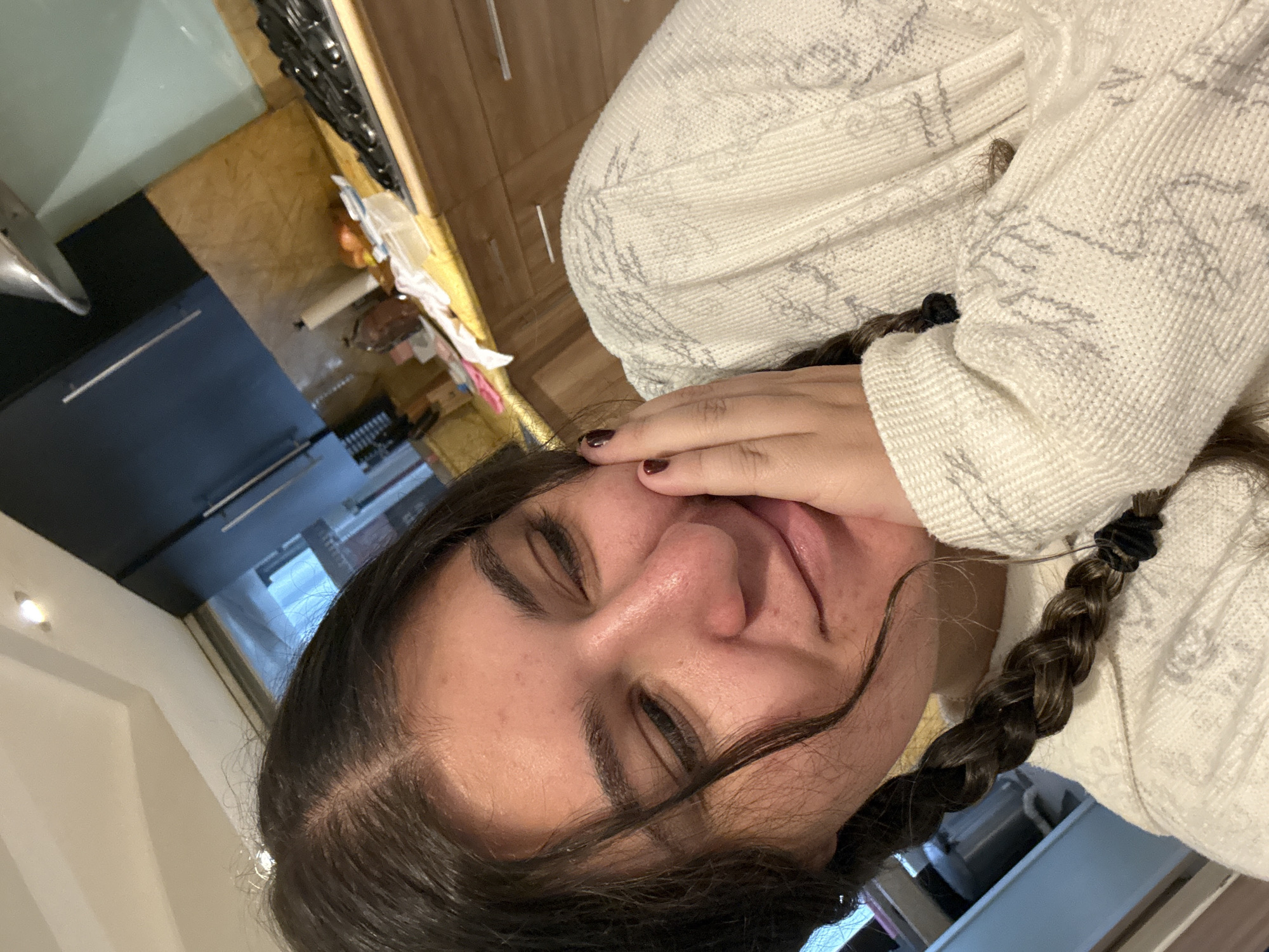 A young woman is depicted in a close-up indoor portrait. She has dark hair styled in braids and is wearing a light-colored, patterned sweater. Her eyes are closed, and her hand is touching her face. The background features a kitchen area with wooden cabinets and a window, hinting at an interior domestic setting. The location is unspecified, and there is no information suggesting it is in Mexico City, Mexico.