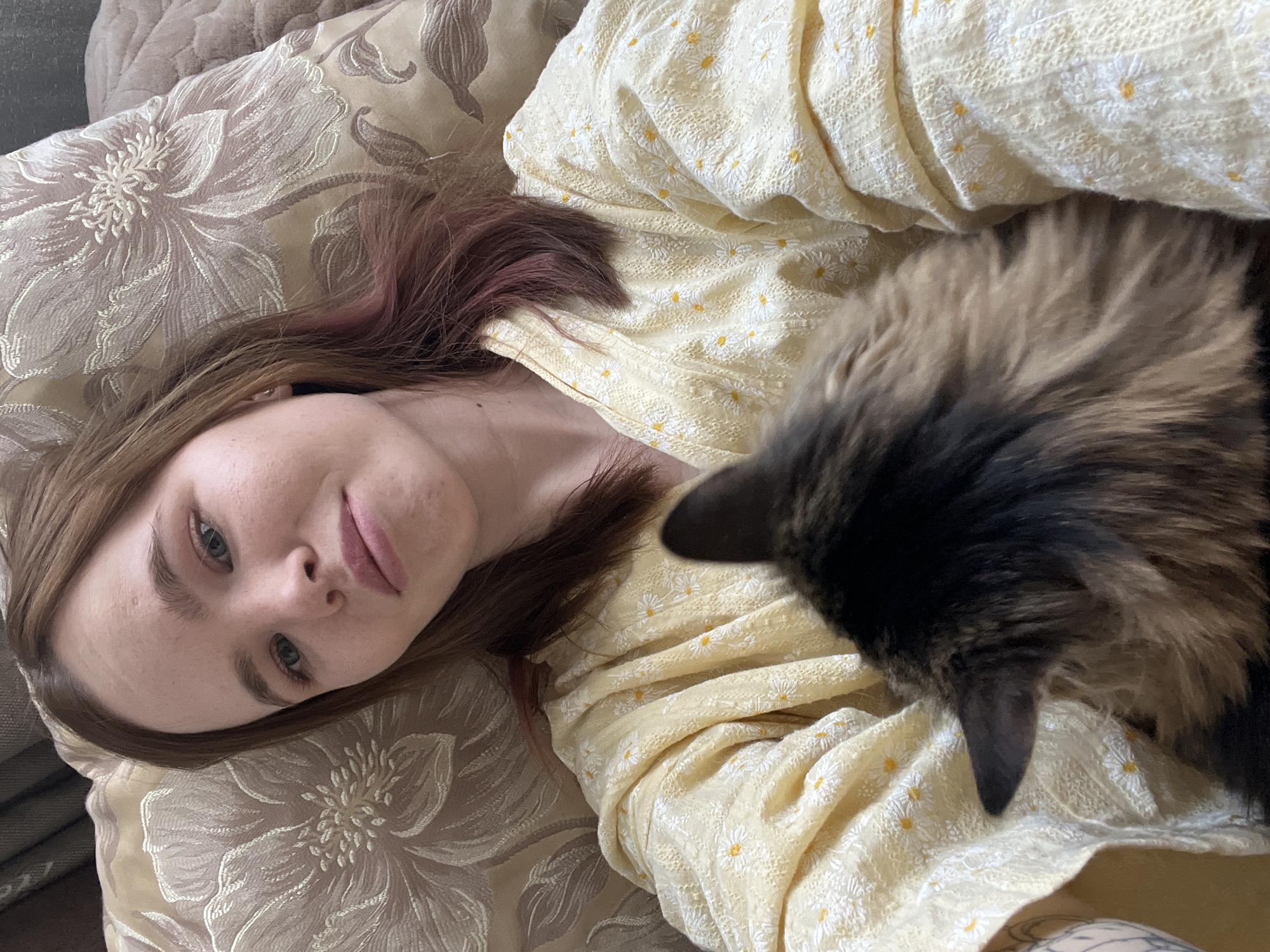 A woman with light skin, light blue eyes, and brown hair with red highlights is lying on a floral patterned pillow. She is wearing a yellow shirt with a small white floral pattern. A brown and black cat is lying on her. The location is Chaikovsky, Russia.