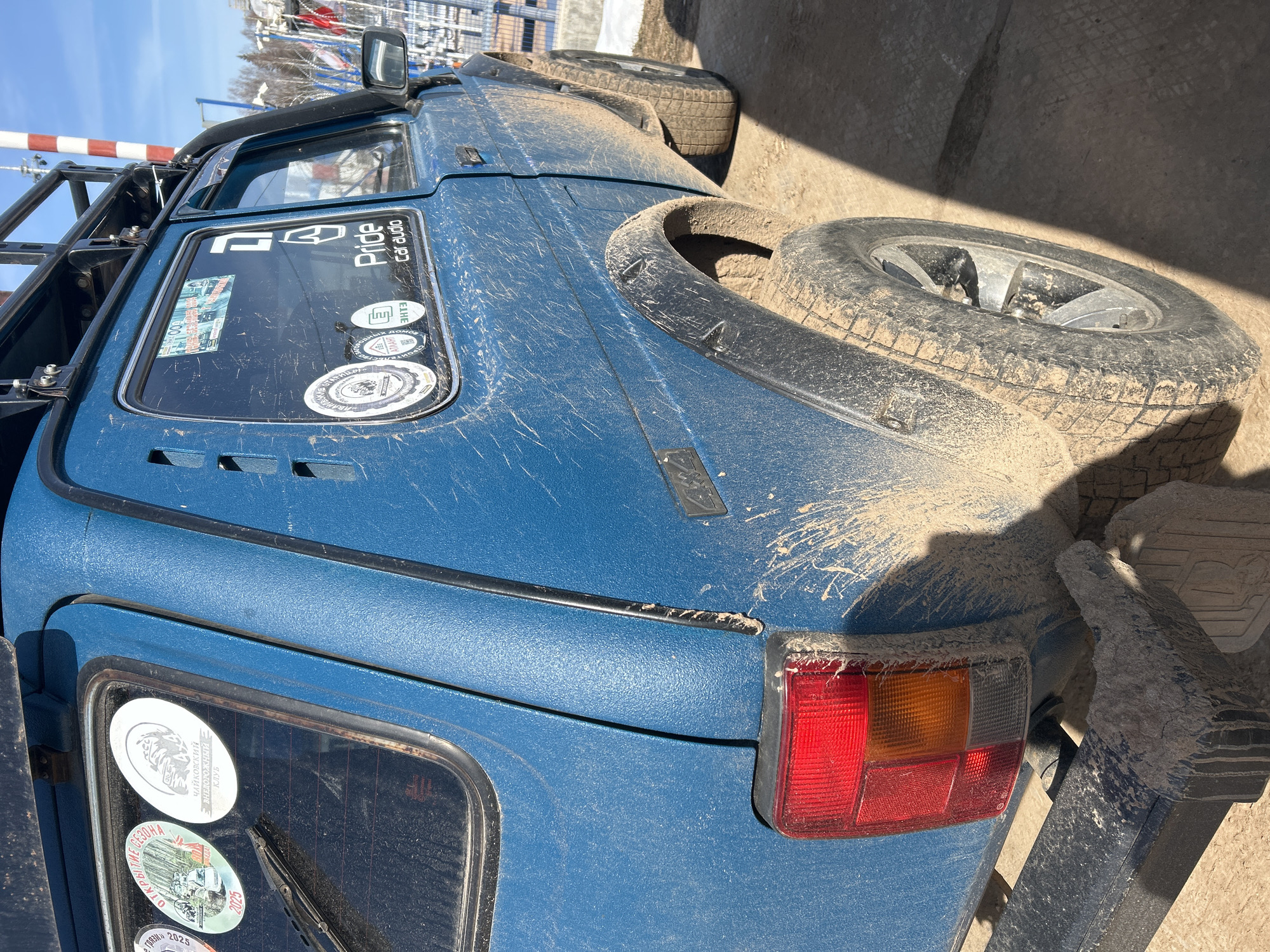 The image captures the rear end of a blue, off-road vehicle covered in mud, suggesting recent use in challenging terrain. A spare tire is mounted on the back. Stickers are affixed to the rear window, one of which says "Pride Car Audio" and a sticker showing "ЧАЙКОВСКИЙ внедорожный клуб" which means "Chaikovskii Off-road Club" . The tail light is a rectangular unit with a red and orange section. The location cannot be determined to be Ural'skoe, Russia, based on this image alone. The presence of a license plate or distinct city landmark to confirm location would be necessary.