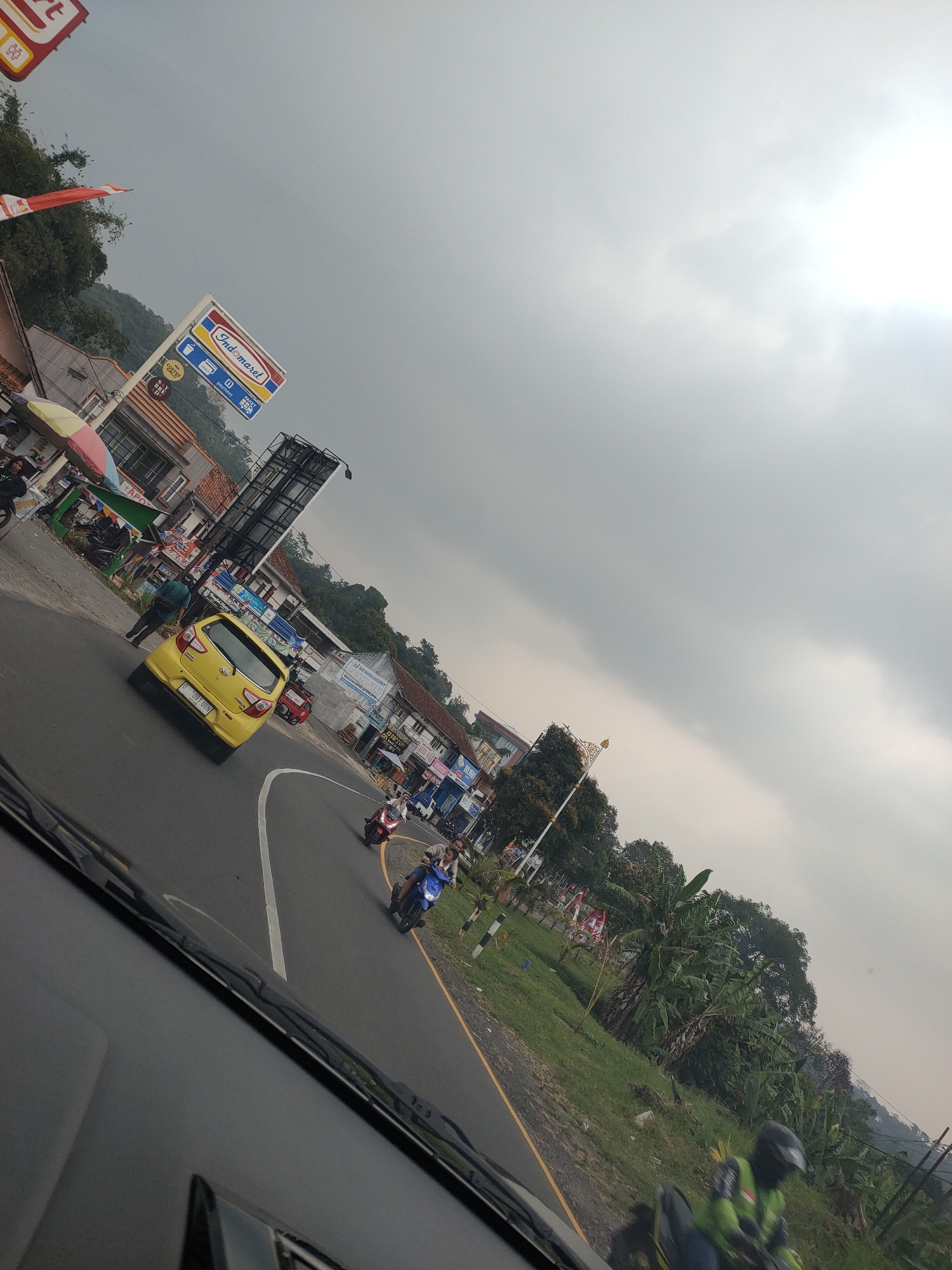 The image, taken from inside a vehicle, shows a street scene in what appears to be a rural or semi-urban area in Subang, Indonesia. The sky is overcast and hazy, with the sun faintly visible through the clouds on the right side. The road curves to the left, with a yellow car in the foreground and several motorcycles following behind. Buildings line the right side of the road, including a store with an "Indomaret" sign, suggesting it's a convenience store. There are also trees and lush vegetation on the left side of the road, indicating a natural environment. The overall impression is one of a typical day in a developing area, with a mix of commercial activity and natural landscapes.