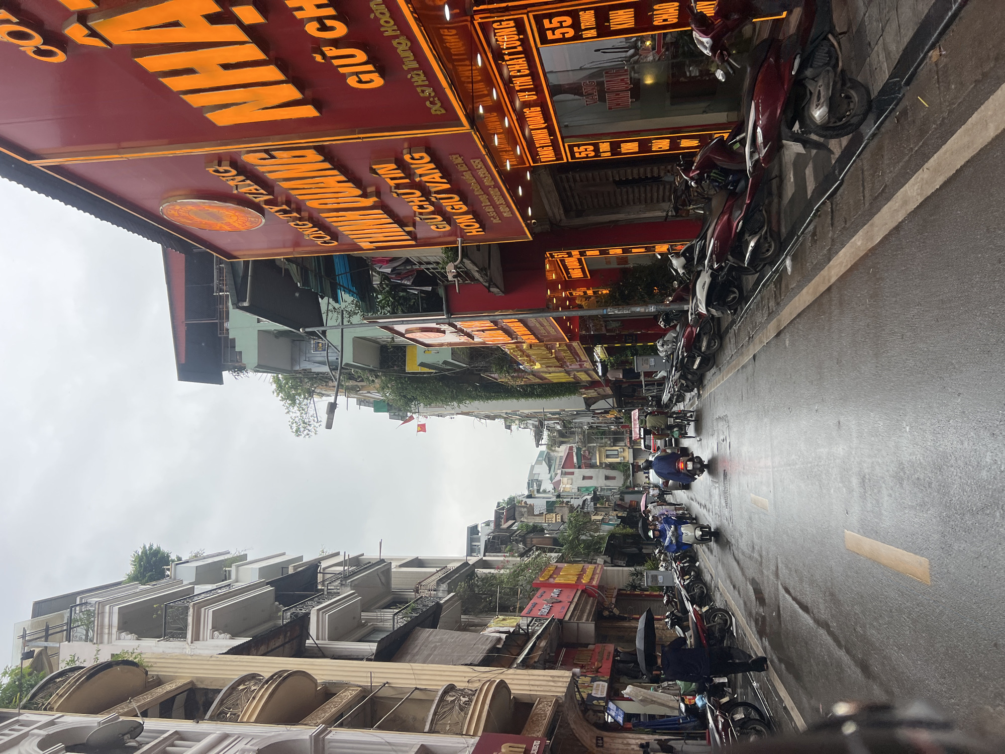 The image depicts a street scene in Hanoi, Vietnam, likely on a rainy day, as indicated by the wet pavement and people holding umbrellas. The street is lined with multi-story buildings, many of which house businesses with prominent signage, often illuminated with neon lights. These businesses appear to be gold and jewelry stores, with names like "THỊNH QUANG" and "VÀNG" (gold) visible. The architecture of the buildings is typical of Vietnamese cities, with narrow facades and balconies on upper floors.
Several motorbikes and scooters are parked along the sides of the street and are also in motion. People are seen riding these vehicles, some wearing rain gear, and at least one person is walking with an umbrella. A man is seen walking away from the camera, holding a black umbrella. Another person on a scooter is facing away from the camera, wearing a blue rain jacket. A person on a bicycle with a passenger is visible further down the street. The road itself has painted lane markers, and there are puddles of water on the surface. The sky is overcast, contributing to the dim lighting. The overall impression is a bustling urban environment with commerce and daily life continuing despite the weather.