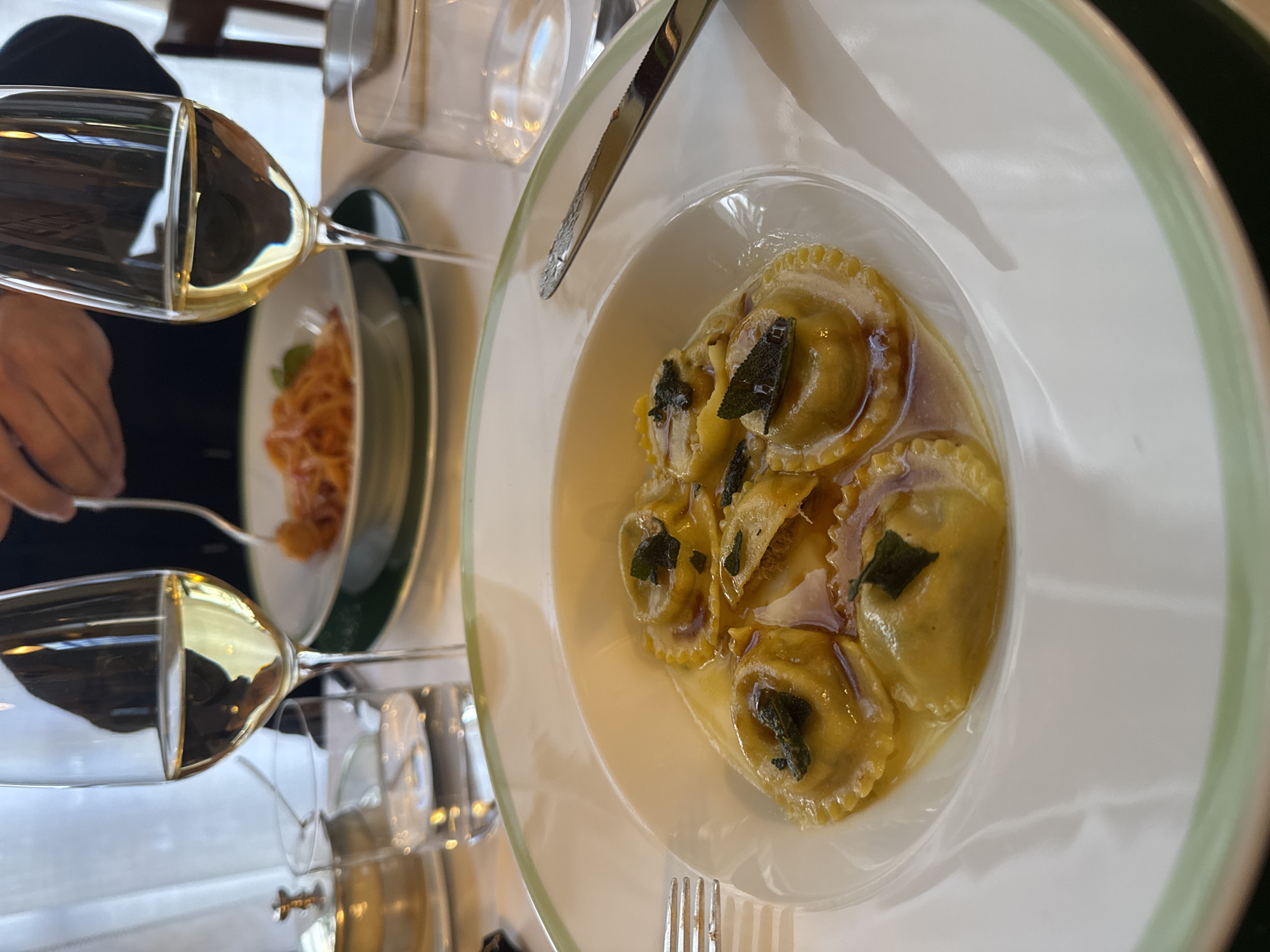 The image captures an indoor dining setting in Milan, Italy.
The foreground presents a white ceramic bowl, bordered by a light green rim, containing seven round ravioli in a translucent, light brown sauce, each garnished with a dark green herb, likely sage. A silver four-tine fork is partially visible on the left, and a serrated silver knife on the right, flanking the bowl. Behind the foreground dish, a clear stemware glass holds a pale yellow liquid, presumably white wine, positioned next to a tumbler containing clear liquid, likely water.
In the midground, a second diner's meal is visible: a white ceramic plate, also with a light green rim, holding a portion of orange-red pasta. A person's hands are shown engaging with this pasta using a silver fork. Another identical stemware glass with pale yellow liquid is present near the pasta dish.
The table surface is covered by a white tablecloth. A small, lidded silver vessel is discernible on the left side of the table. The background shows white, draped window coverings and a partial view of a dark wooden chair, indicating a restaurant environment.