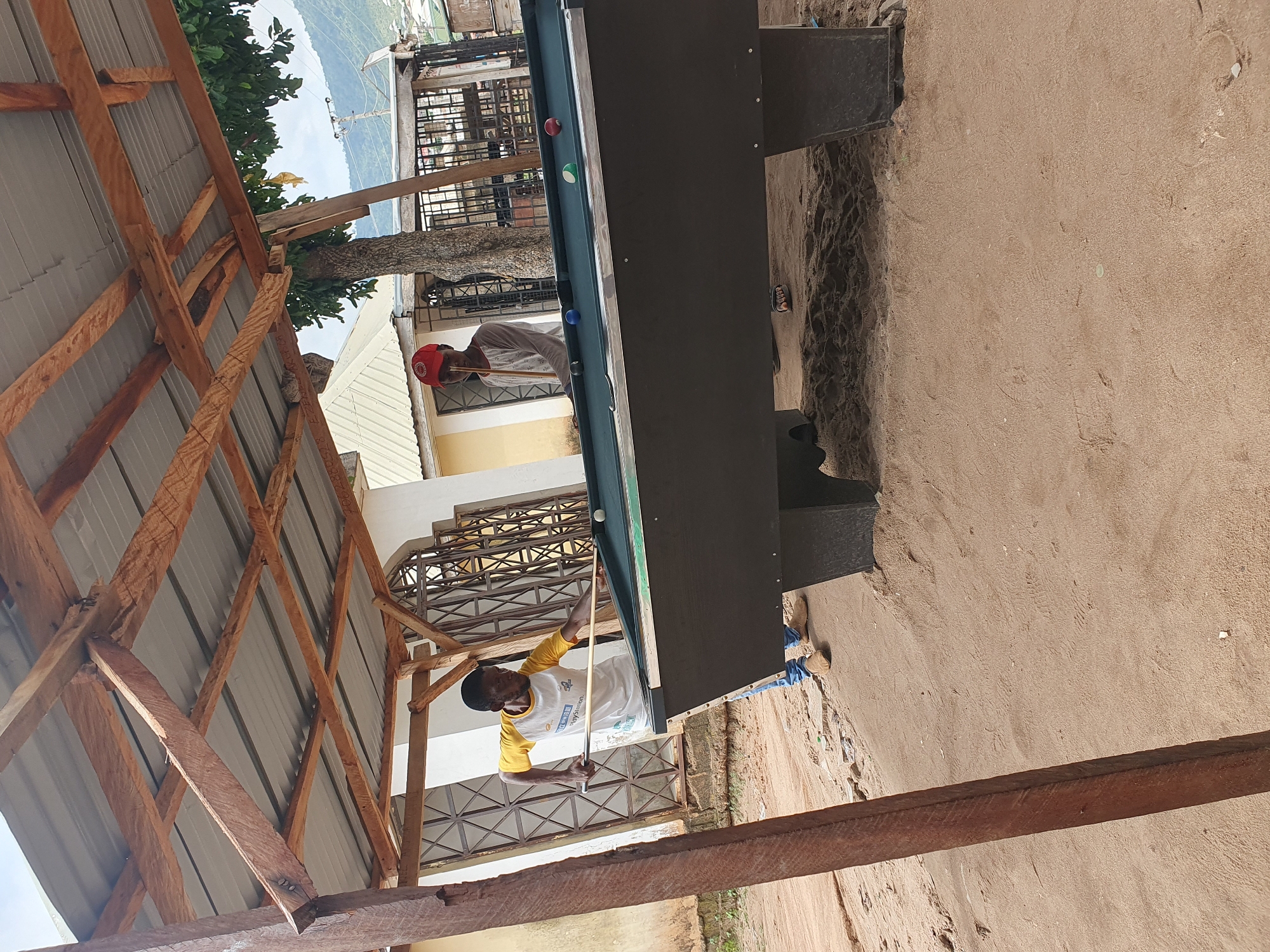 The image depicts a scene in Lokoja, Nigeria. Two men are playing pool outdoors. One man, wearing a yellow and white t-shirt, is taking a shot with a pool cue. The other man is wearing a red cap. The pool table is situated on a dirt surface, near a building with decorative metalwork on its windows. An open-sided wooden structure with a corrugated metal roof covers part of the scene. In the background, trees and distant buildings can be seen.