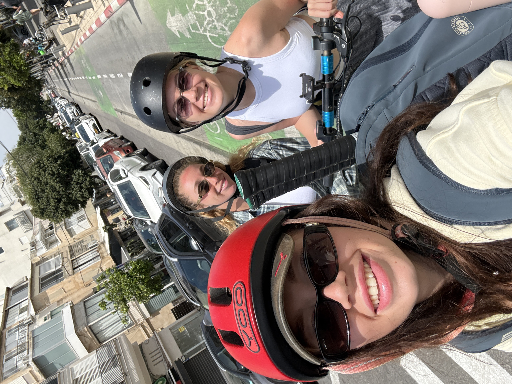 Three women on scooters pose for a selfie on a street in Tel Aviv, Israel. The first woman, positioned in the foreground, wears a red helmet and sunglasses. The second woman, in the middle ground, also wears a helmet and sunglasses. The third woman, furthest from the camera, wears a black helmet and sunglasses. All three are smiling. A green bike lane runs parallel to parked cars on the left side of the frame. Buildings are visible across the street.